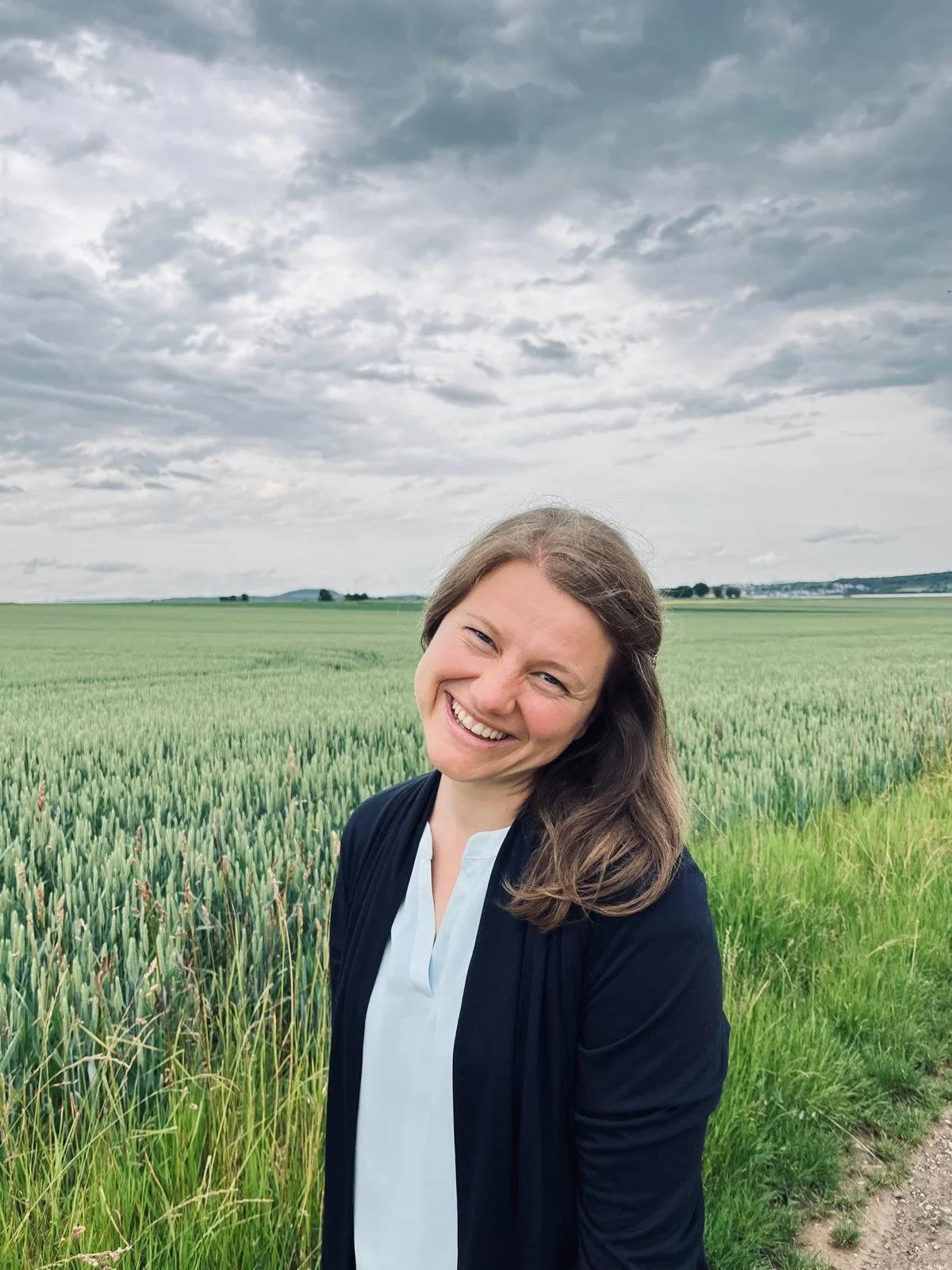 Eine lachende Frau in dunklem Blazer und weißem Hemd steht in einer grünen Feldlandschaft mit bewölktem Himmel im Hintergrund.