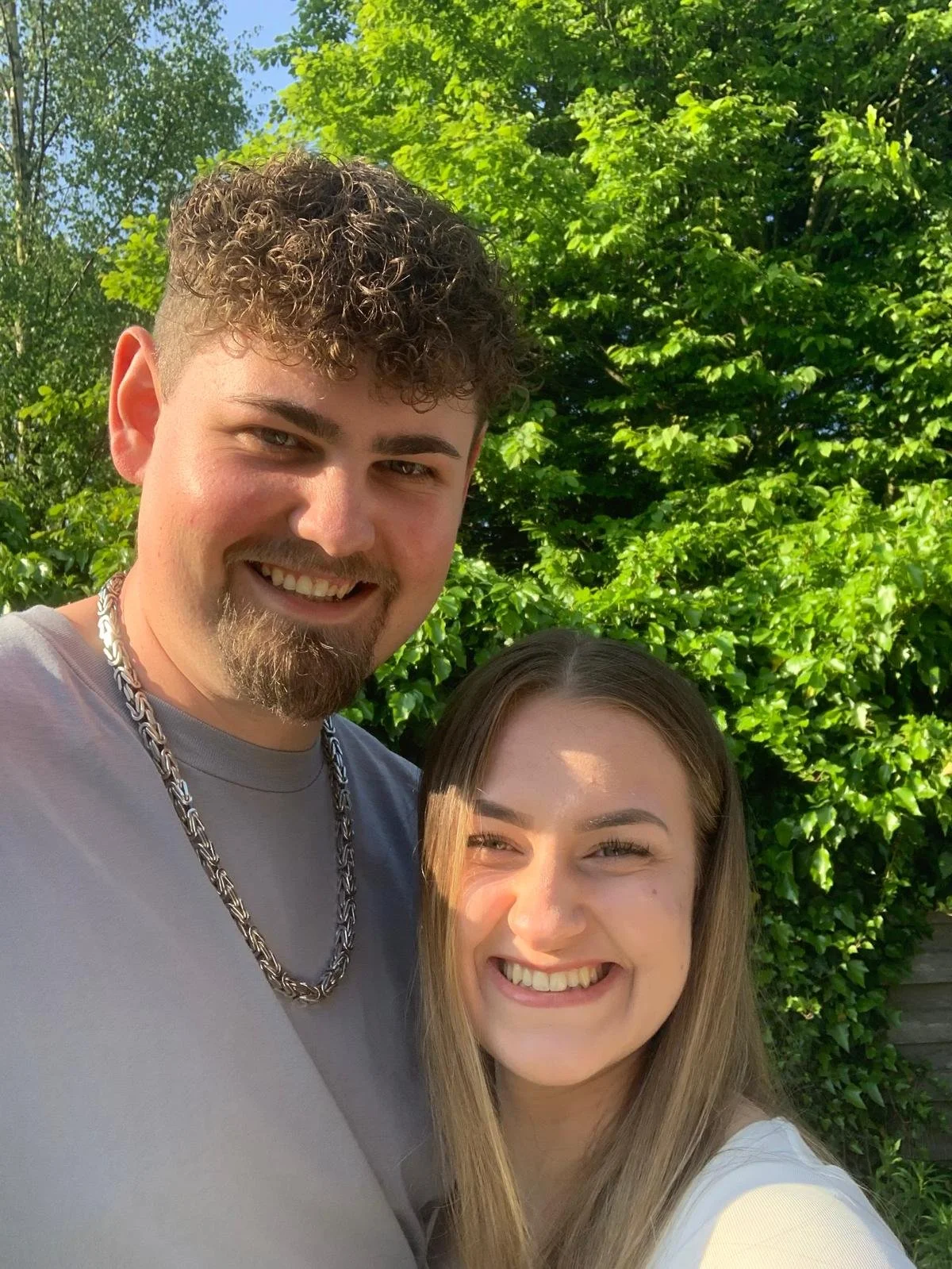 Ein Mann und eine Frau lächeln, stehen vor grünen Büschen an einem sonnigen Tag.