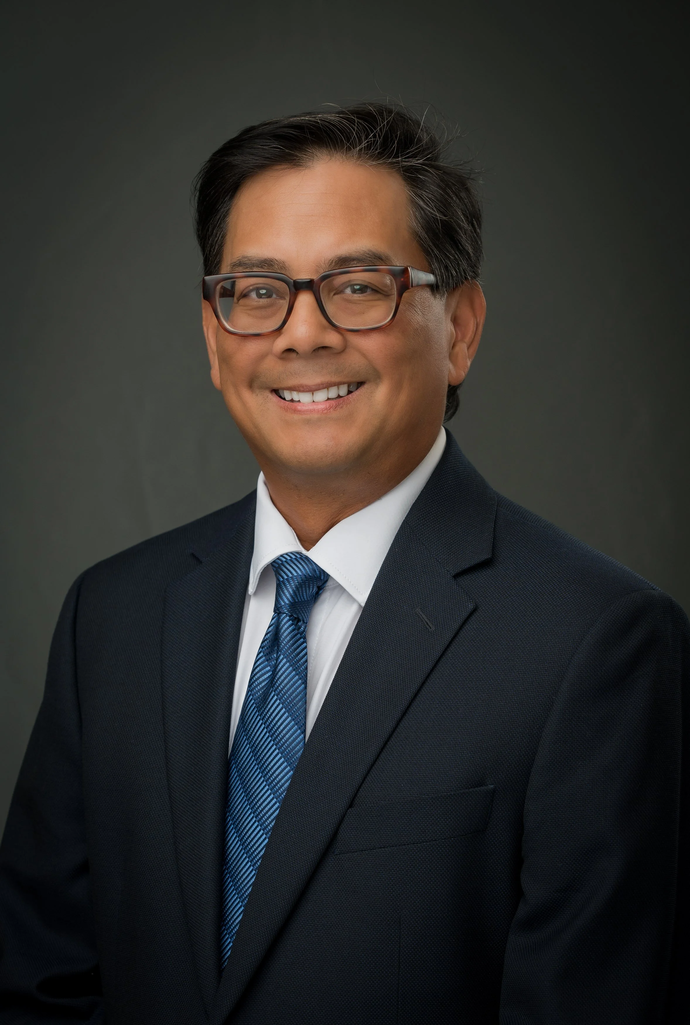 Professional headshot of a man with glasses, wearing a dark suit, white shirt, and blue tie, smiling against a dark background.