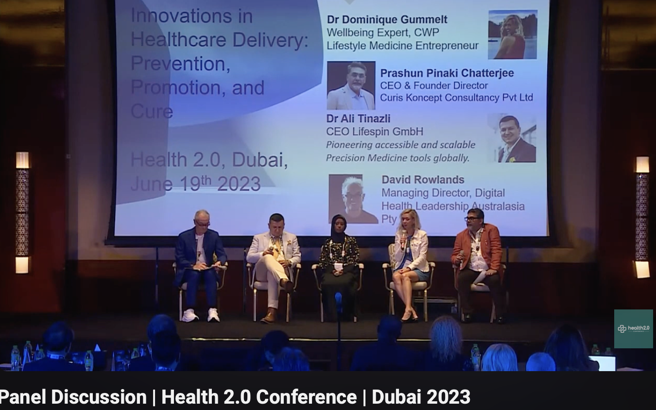 A panel discussion at the Health 2.0 Conference in Dubai 2023, featuring five speakers seated on stage with a large screen behind them displaying information about the event and panelists.
