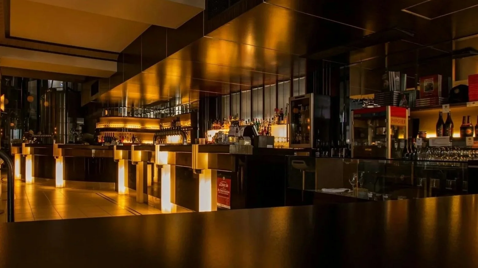 dimly lit bar with golden accents, liquor bottles on shelves, and a dark counter with illuminated bases.