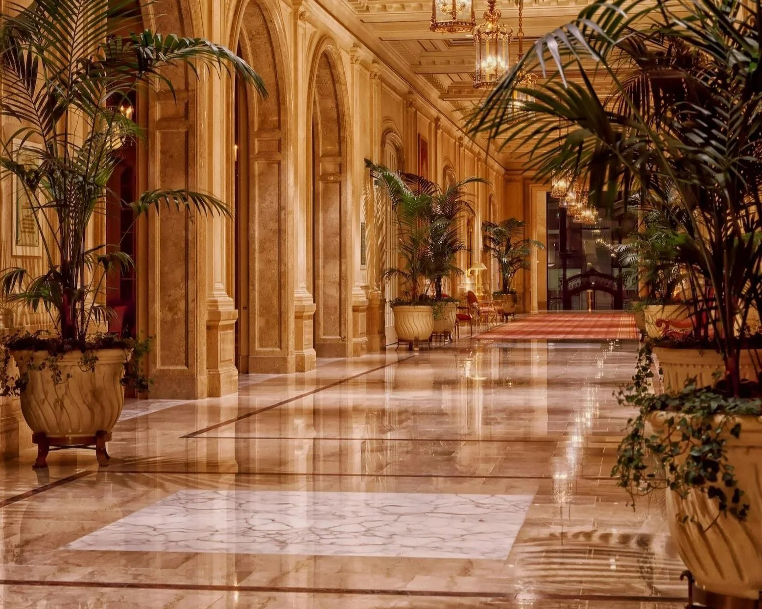 Luxurious hotel or palace hallway with marble floors, ornate golden walls, tall arched mirrors, and large potted plants. Elegant chandeliers hang from a detailed ceiling, and there are plush chairs and a rug at the far end.