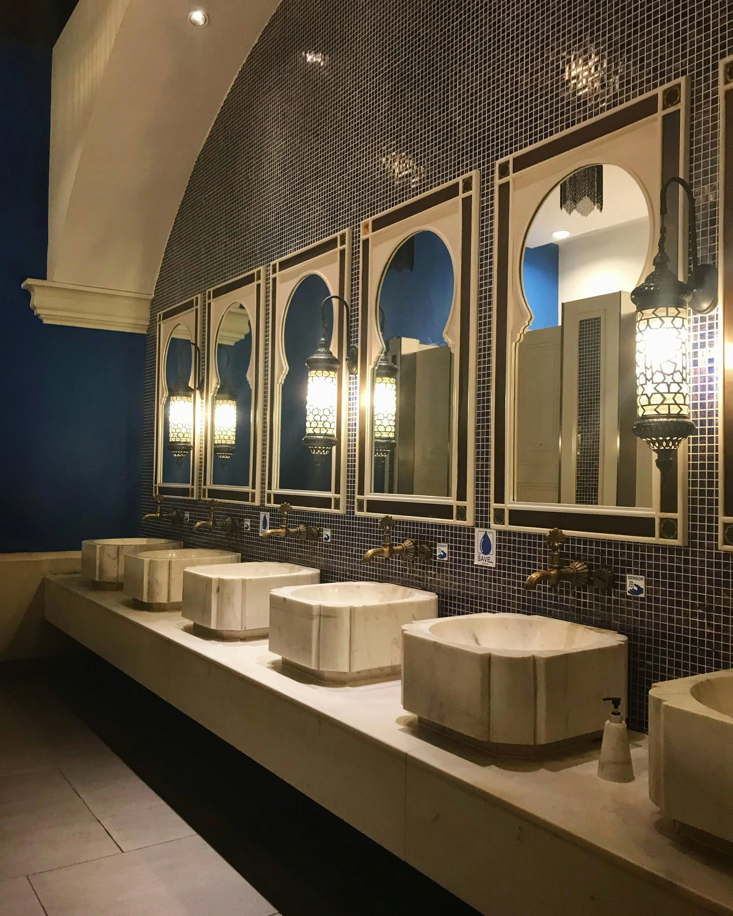 Four marble sinks with gold faucets in front of four mirrors with arched tops and lantern-style lights, in a bathroom with blue walls and small mosaic tile backsplash.