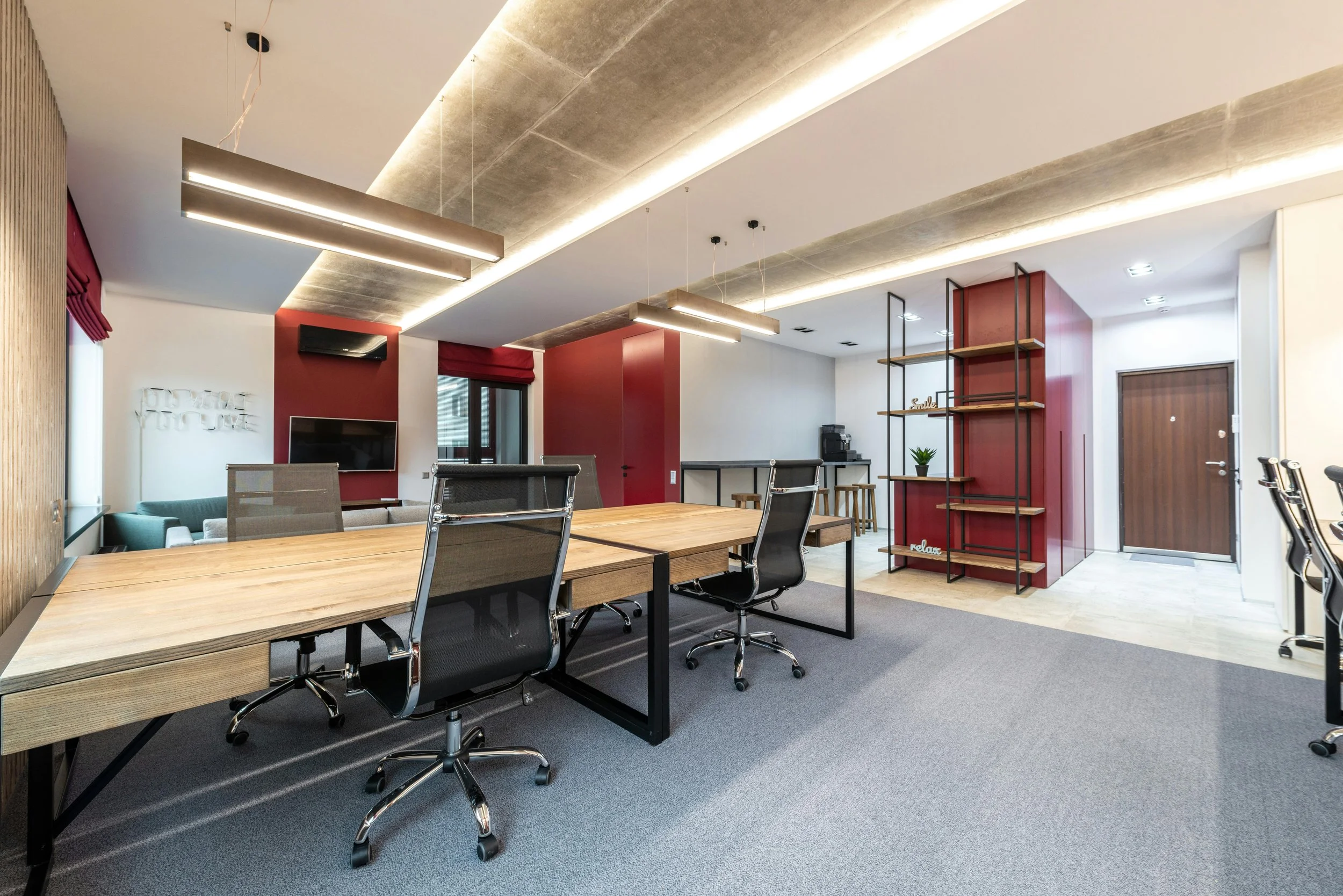 Modern office space with a long wooden table, black office chairs, wall-mounted TV, red accent walls, open shelving with decorative items, and a small coffee station.