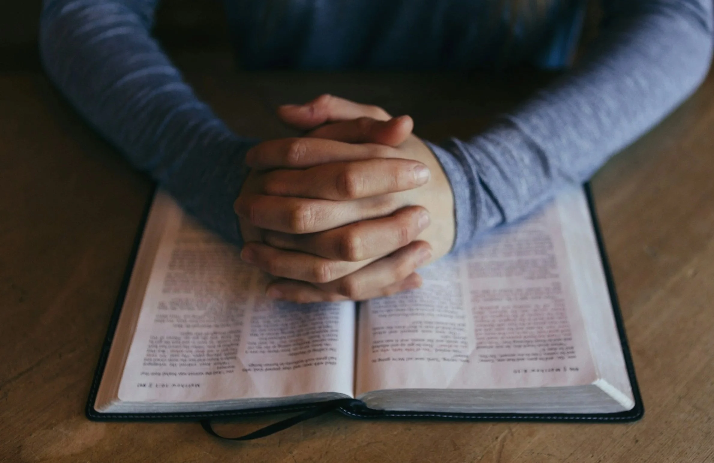 Person praying with hands clasped over an open Bible on a wooden table.