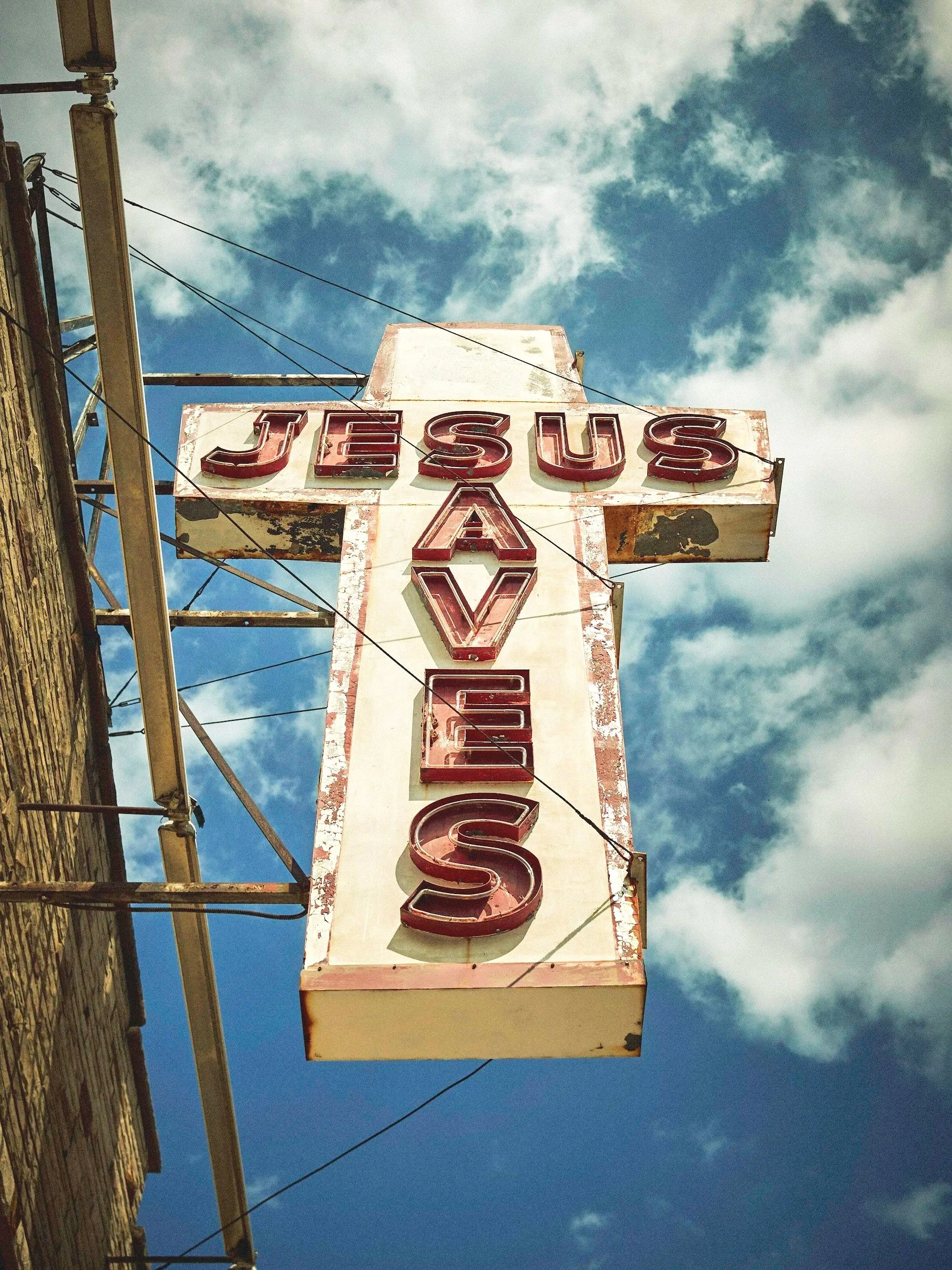 An old vertical neon sign with the word 'JESUS' at the top and 'Saves' vertically below, attached to a brick building against a blue sky with clouds.