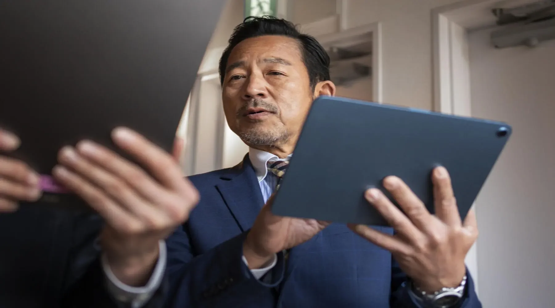 A middle-aged man wearing a blue suit and a striped tie, holding a blue tablet, with a focused expression.