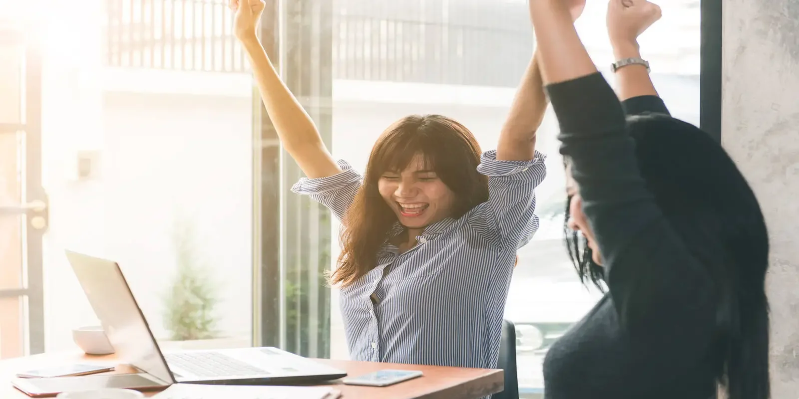 Two women celebrate with raised hands near a laptop in a bright, modern office.