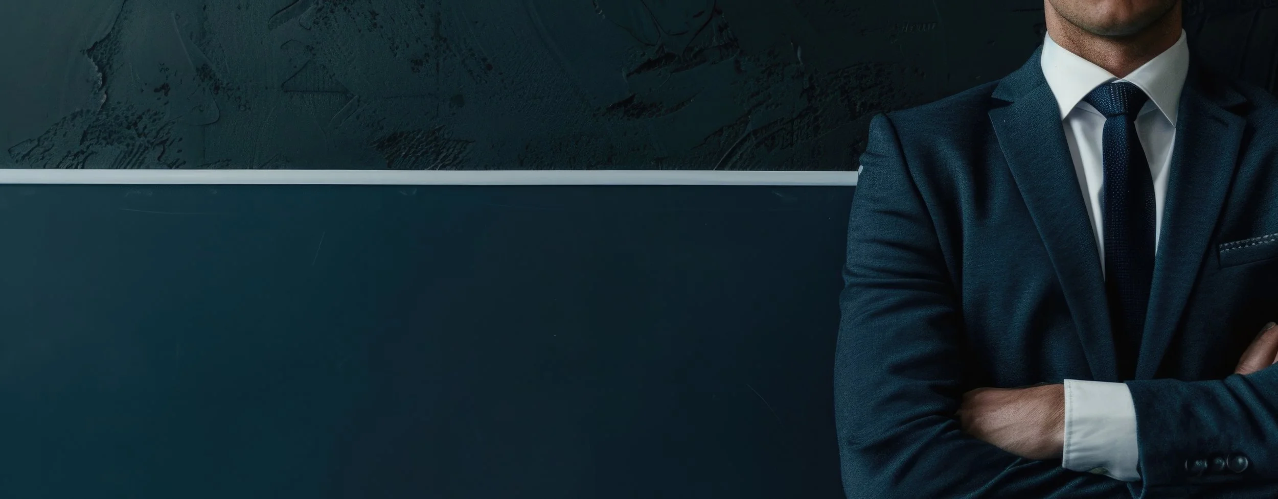 Close-up of a man in a navy blue suit with arms crossed, standing against a dark wall with a textured painting or mural behind him.