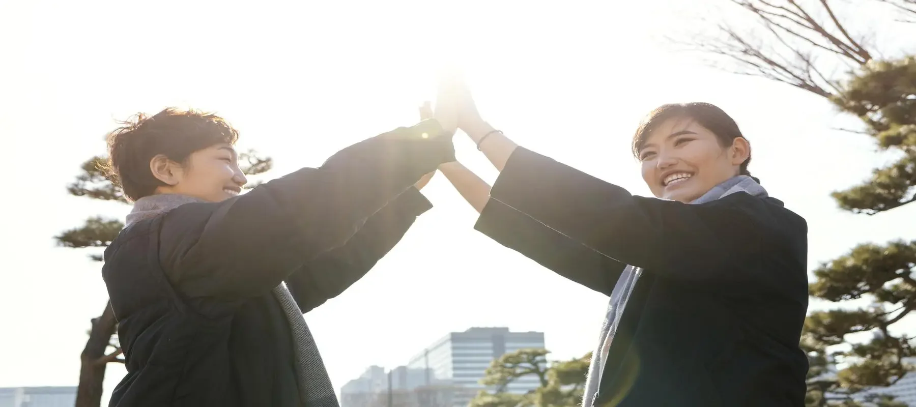 Two smiling women high-fiving outdoors on a bright, sunny day with trees and buildings in the background.