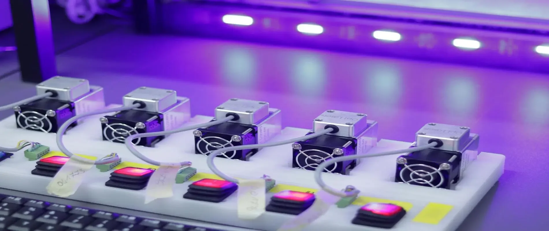 Close-up of a white electronic circuit board with five small fans, wires, and illuminated red and green rectangular components under purple lighting.
