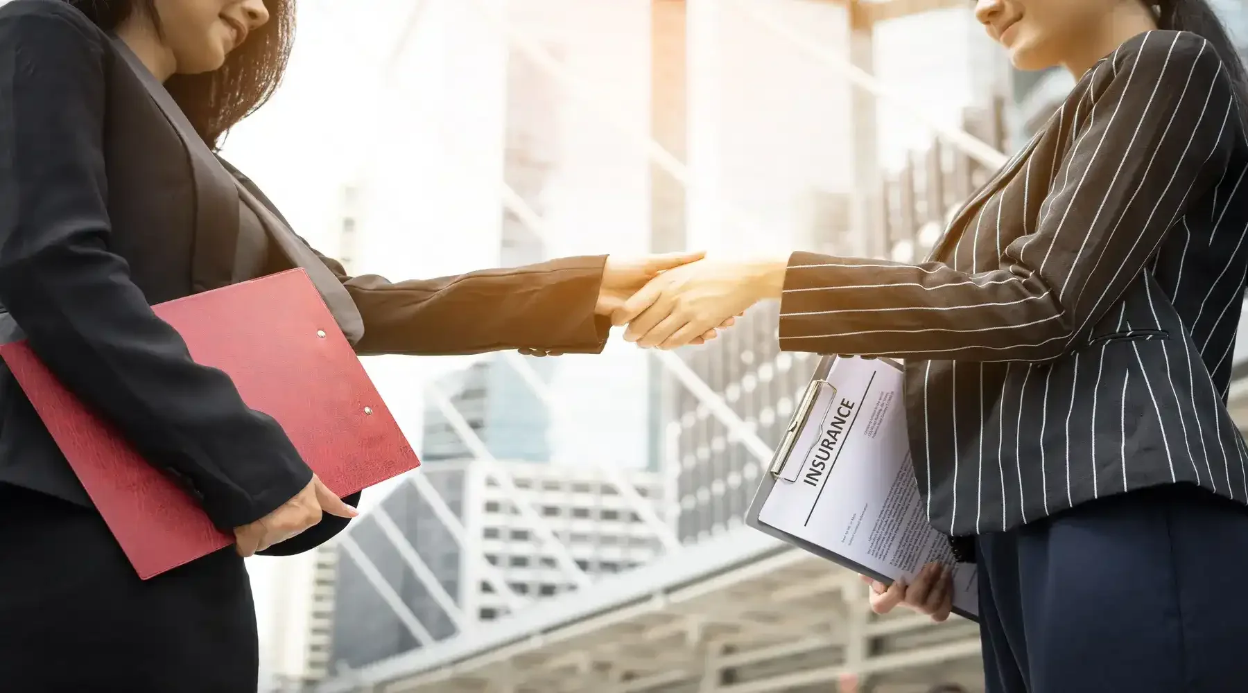 Two businesswomen shaking hands outside in front of a modern building, with one holding a folder and the other holding an insurance document.