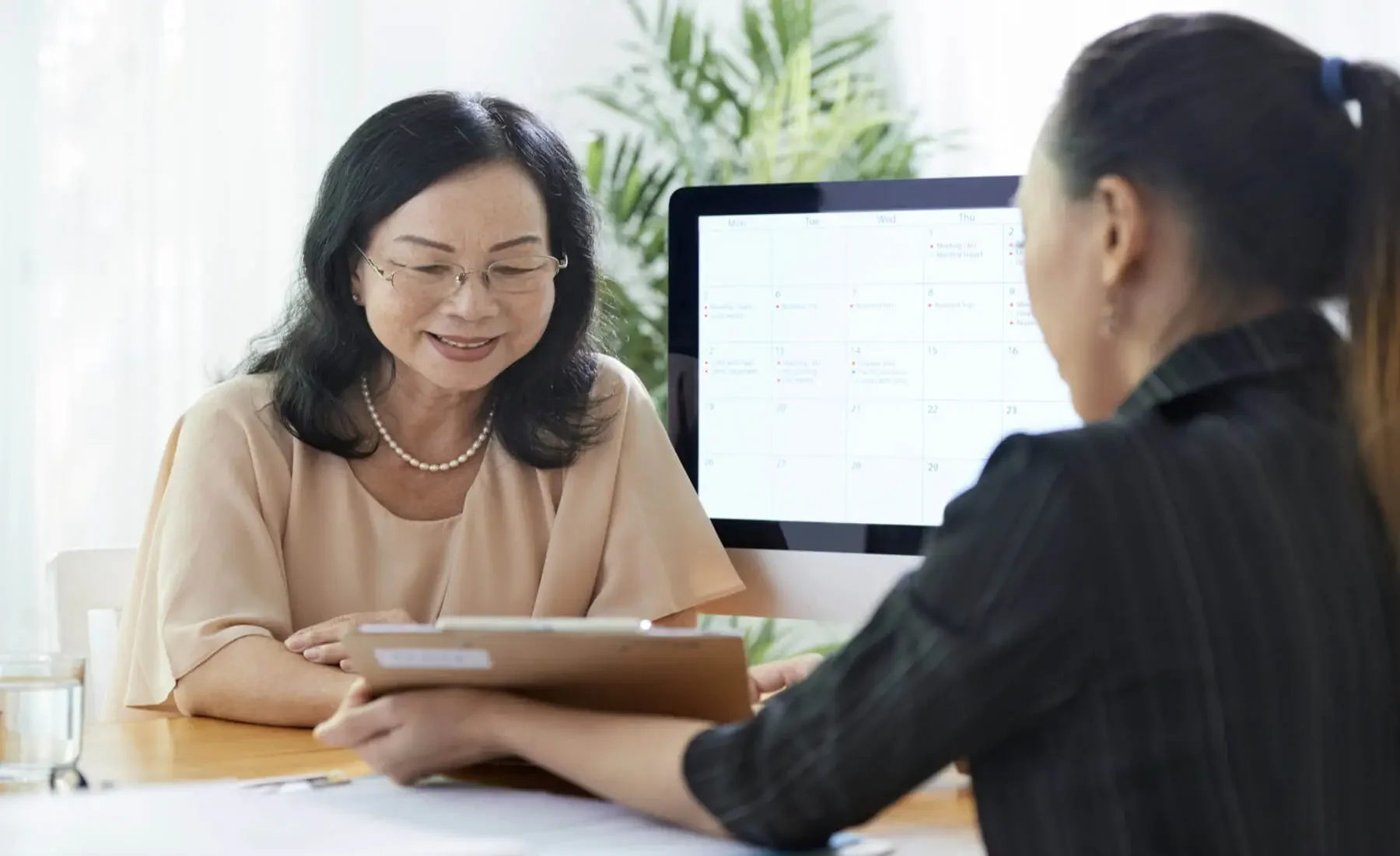 Two women sitting across from each other at a table, one with glasses and a pearl necklace, smiling and holding a folder, the other with a ponytail, reviewing papers, with a computer monitor displaying a calendar in the background.