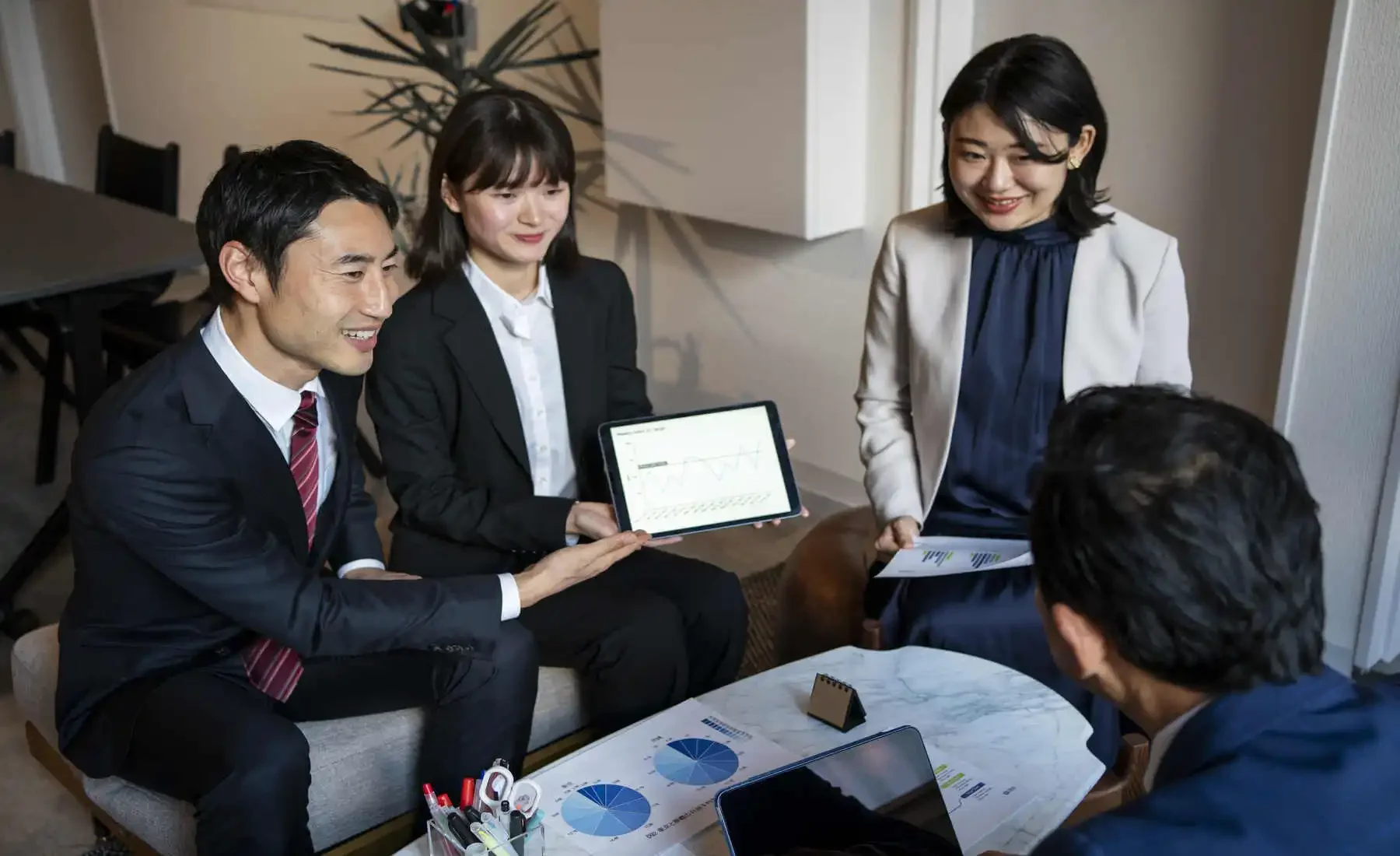 A group of five business professionals in a meeting, with one woman showing a tablet with graphs and charts to the others.
