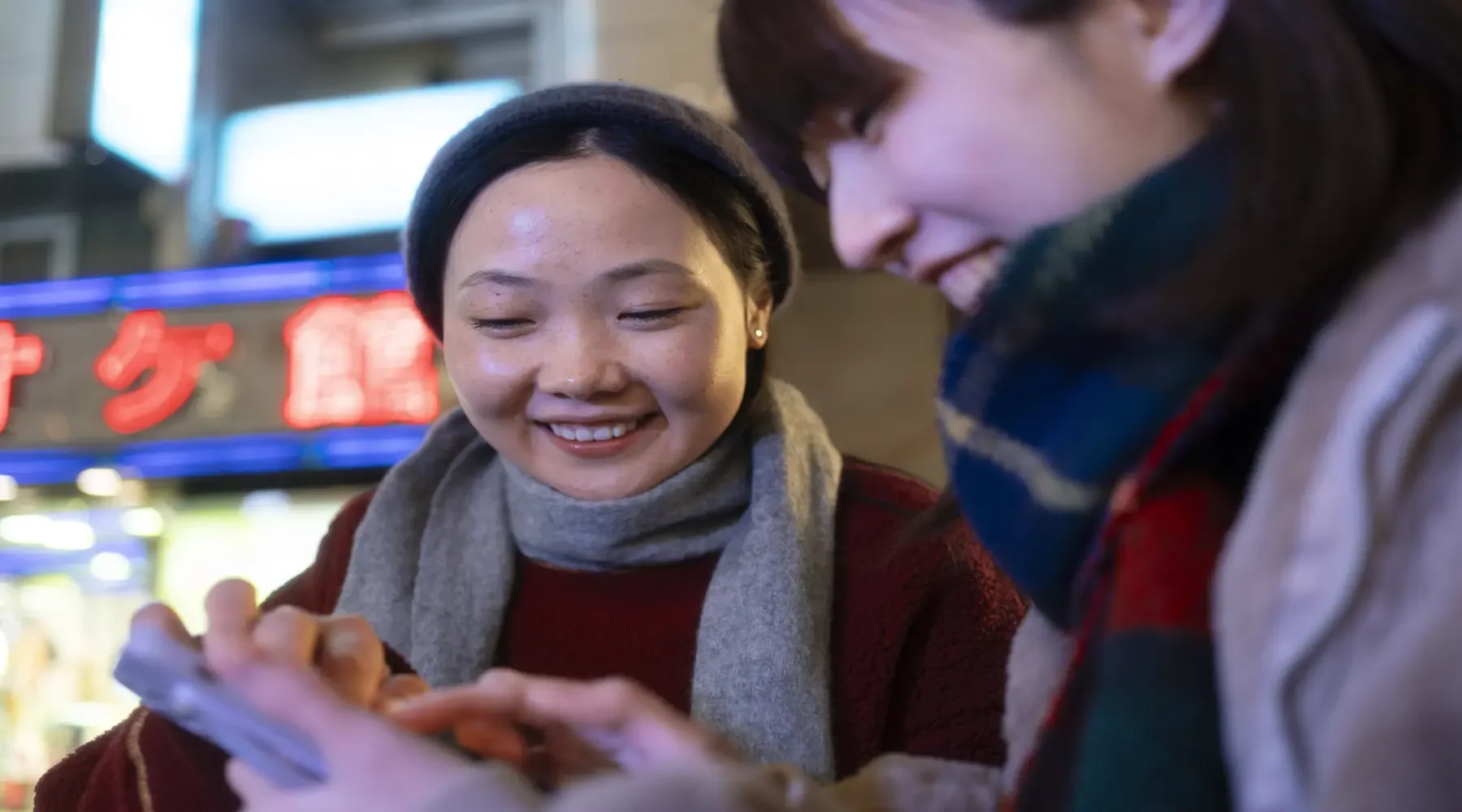 Two women smile and look at a smartphone together on a city street at night, with neon signs in the background.