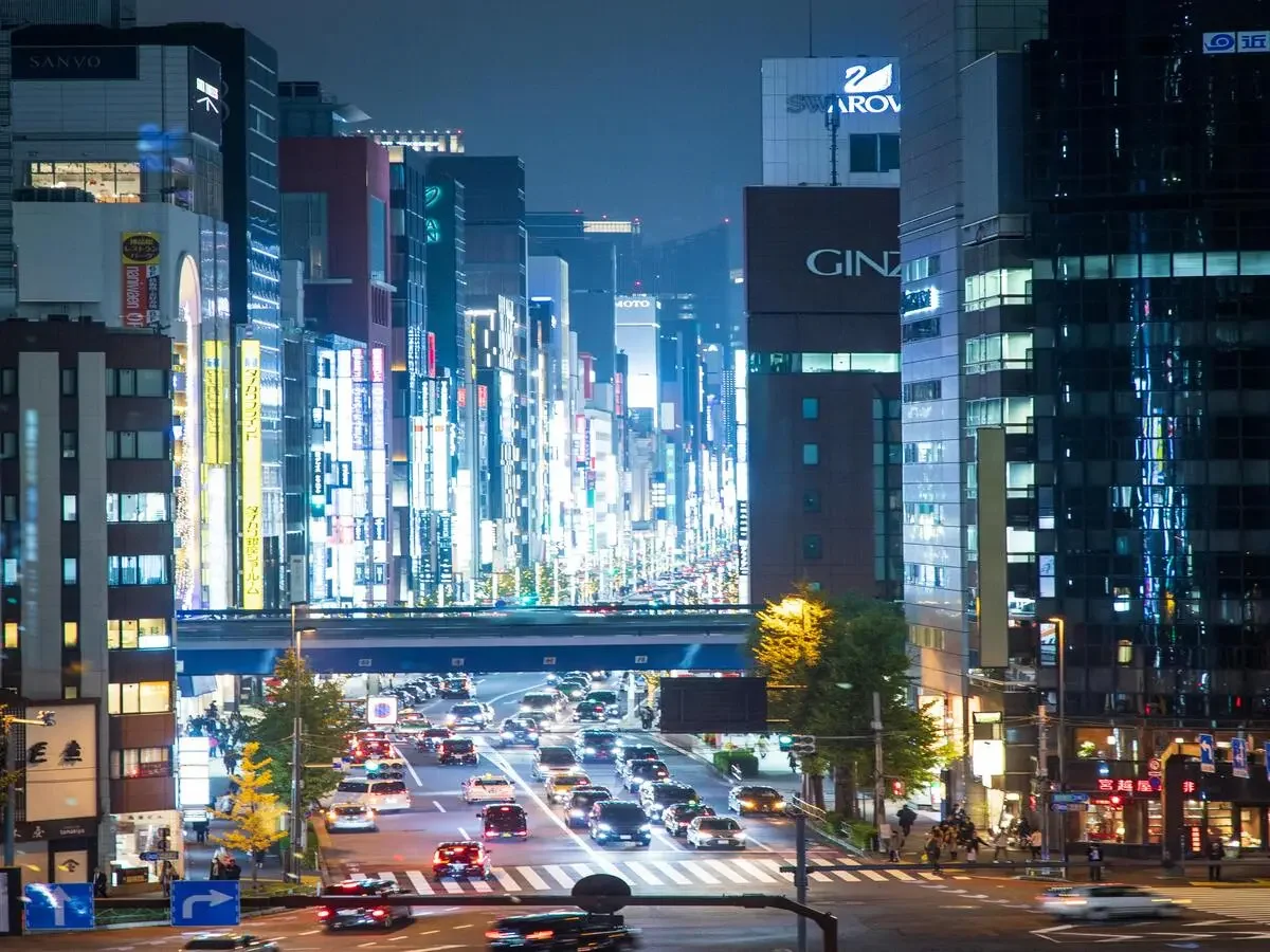 Night view of a busy city street with illuminated modern skyscrapers and commercial buildings, traffic moving through an intersection, and colorful neon signs on the buildings.
