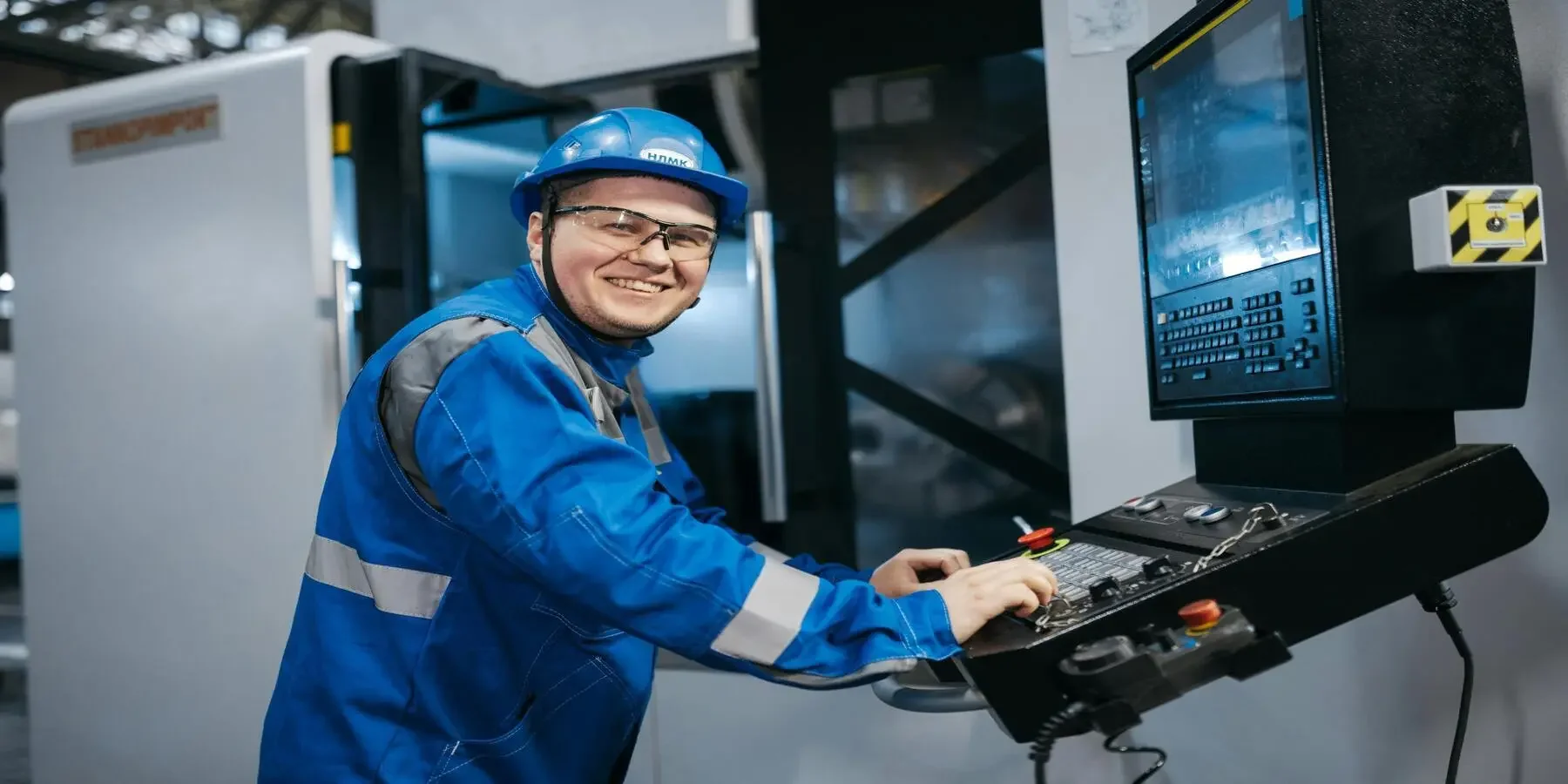 A worker in blue safety gear, including a helmet and glasses, operating a control panel in an industrial setting, smiling at the camera.