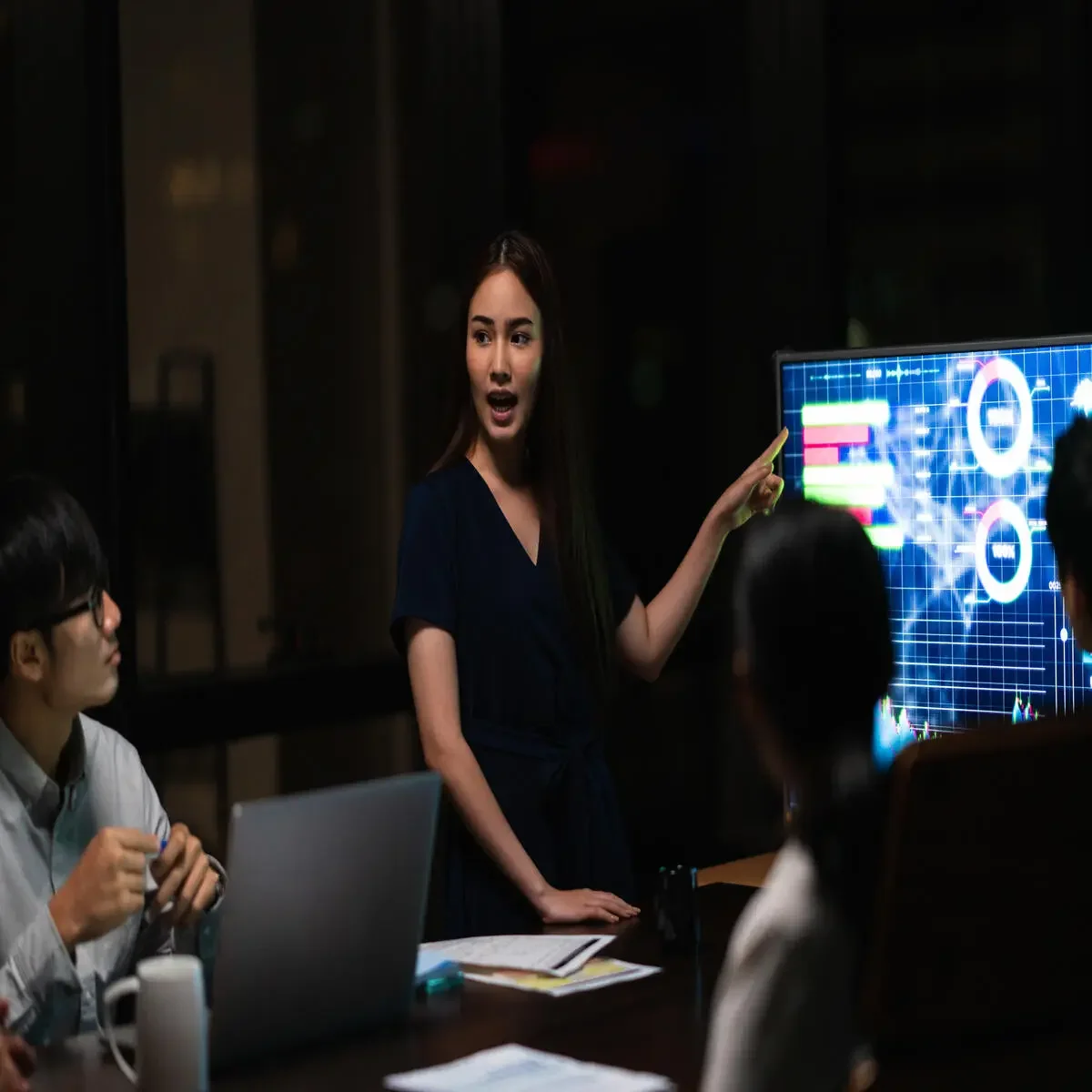 A woman giving a presentation about financial data to a group of people in a dark room, with a large digital screen displaying graphs and charts.