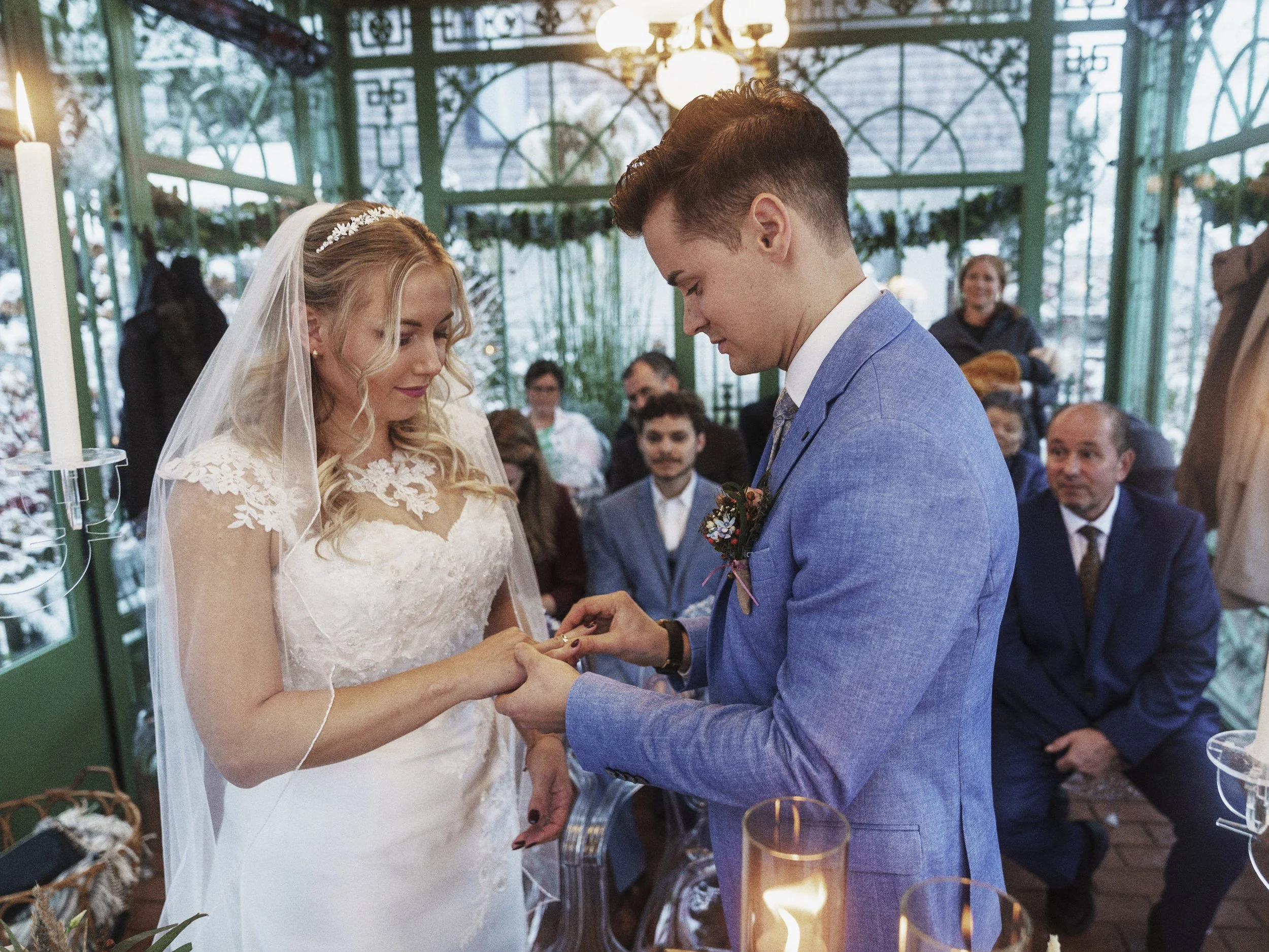 Ein Brautpaar bei der Trauung, der Bräutigam steckt der Braut einen Ring an, im Hintergrund sitzen Gäste in einer grünen Gartenlaube.