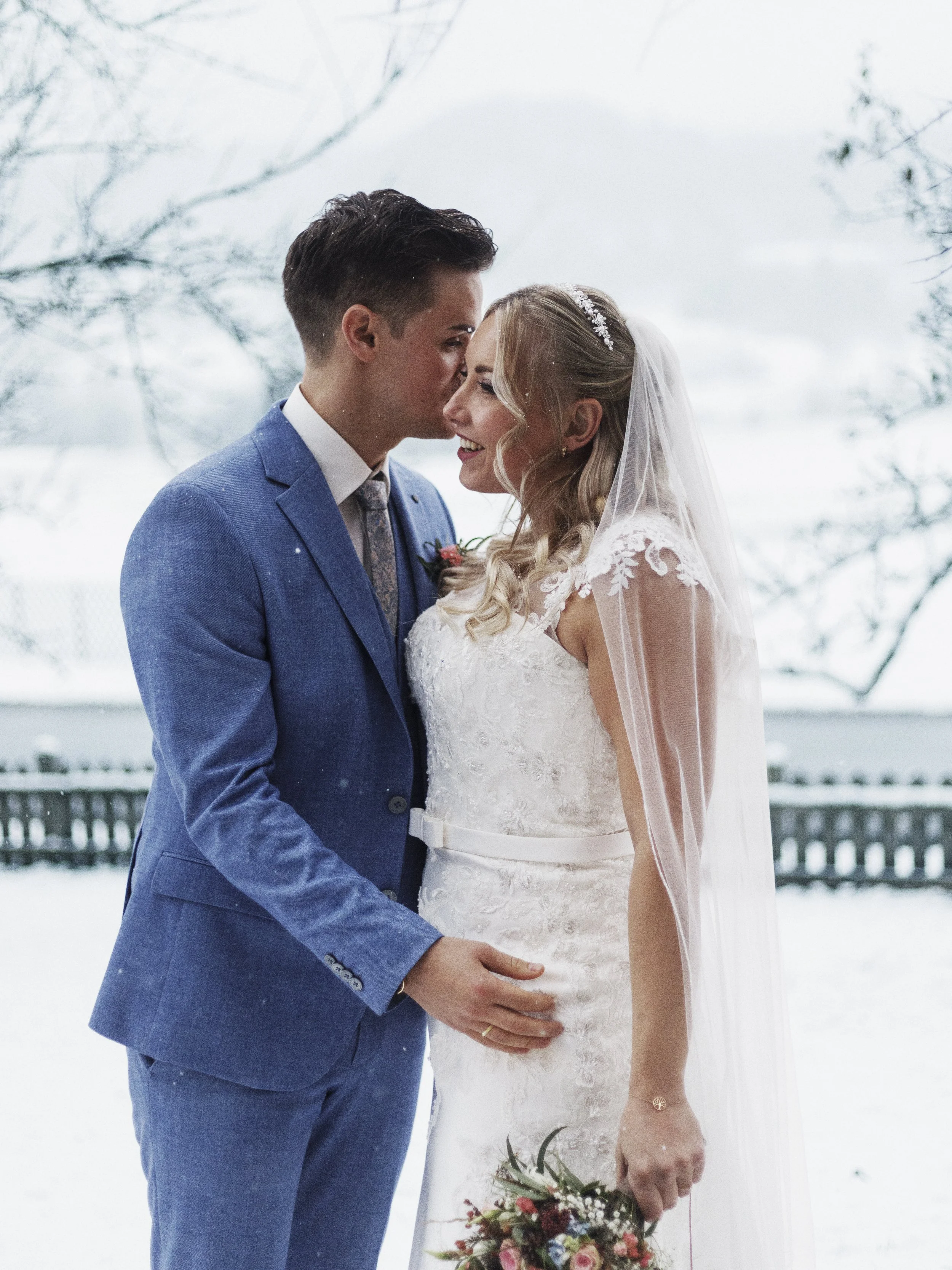 Ein Brautpaar, das bei Schneewetter draußen steht, mit Bäumen im Hintergrund, der Bräutigam küsst die Braut auf die Stirn, sie trägt ein weißes Hochzeitskleid mit Schleier, er trägt einen blauen Anzug.