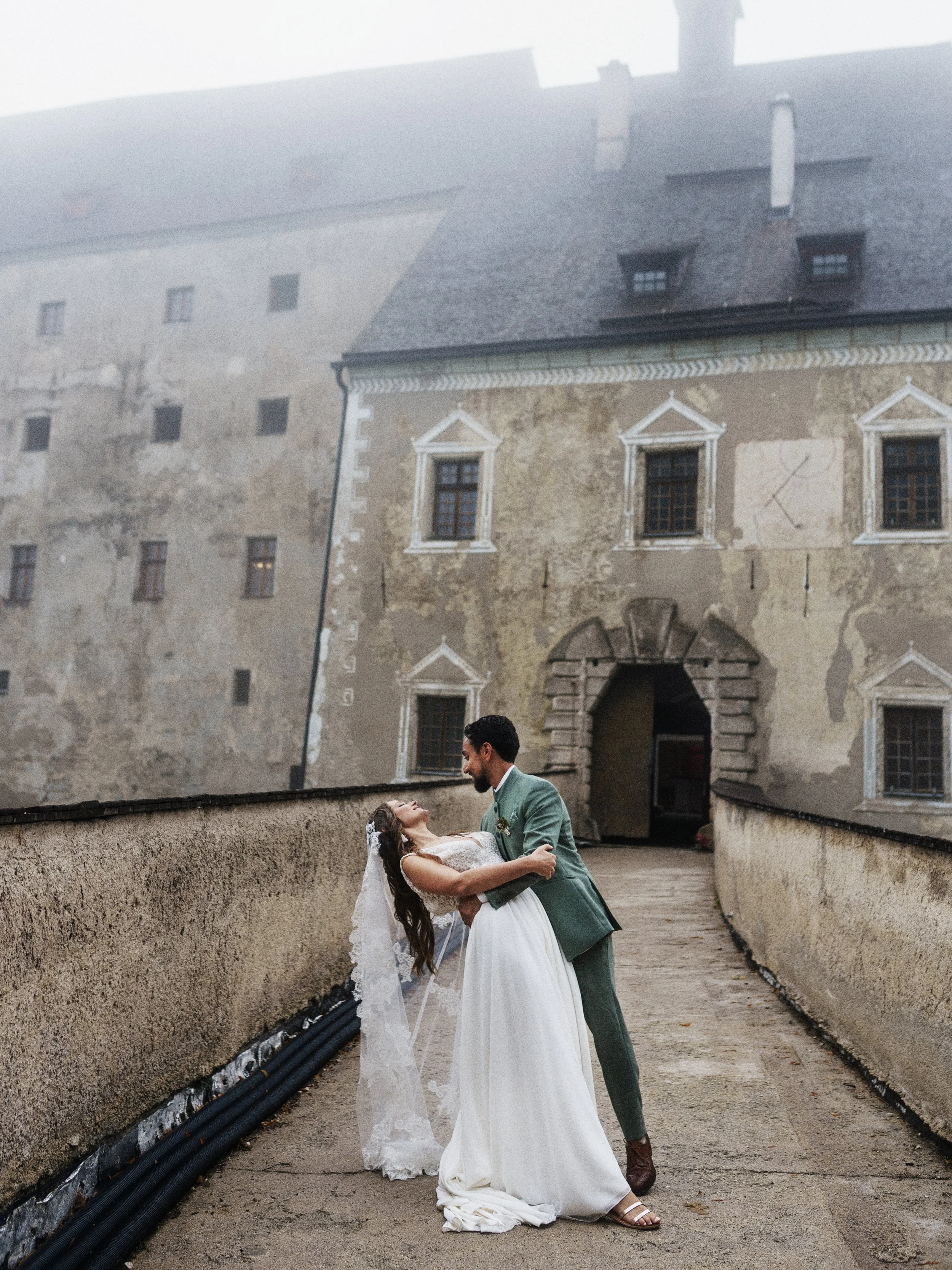Ein Brautpaar tanzt auf einem alten Burgmauer, im Hintergrund ein historisches Gebäude mit Steindach und kleinen Fenstern, nebliger Himmel