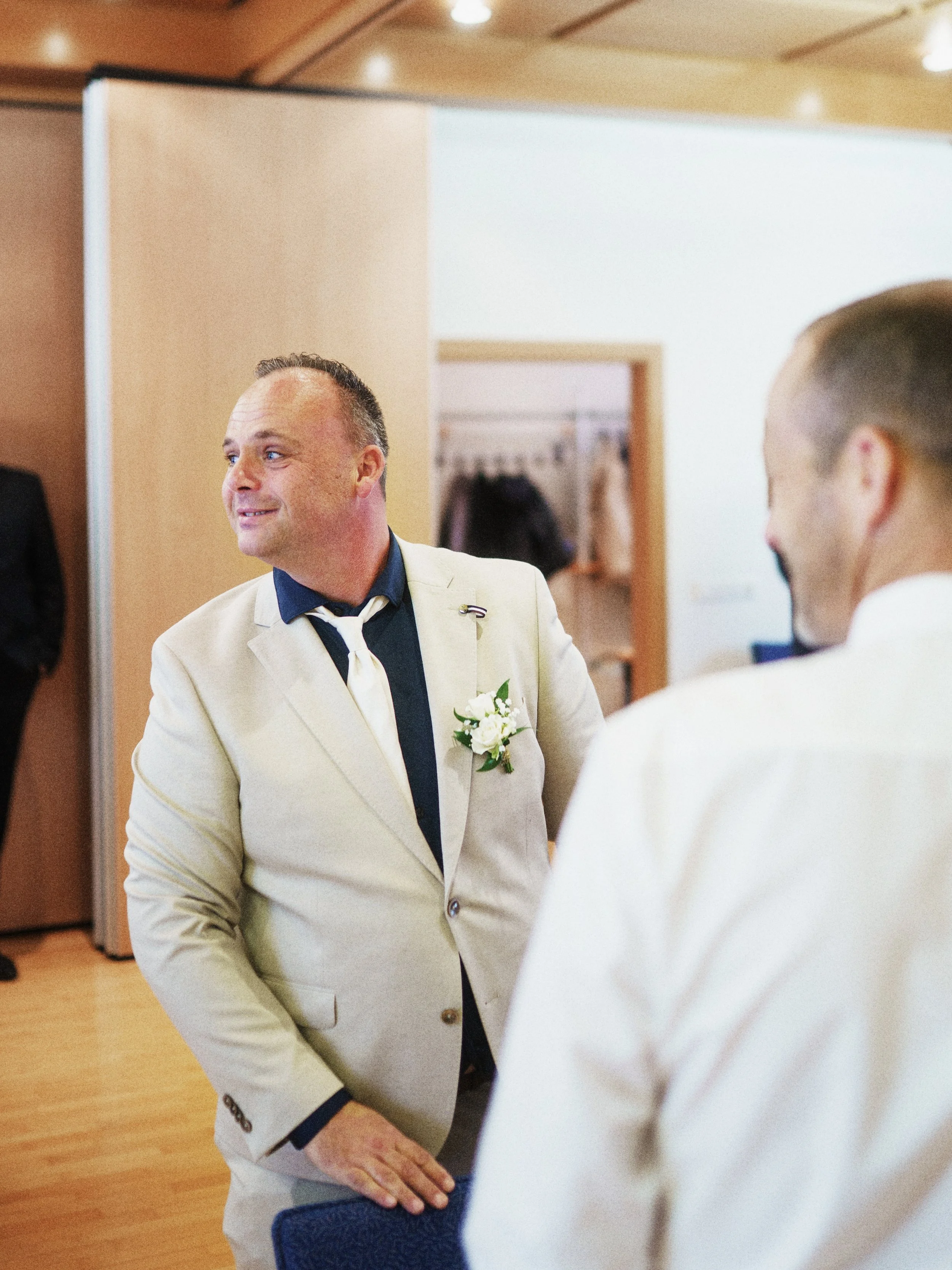 Zwei Männer im Gespräch, einer trägt einen beigen Anzug mit weißer Blume als Boutonnière, der andere trägt ebenfalls einen weißen Anzug. Das Bild wurde wahrscheinlich bei einer Hochzeitsfeier aufgenommen.