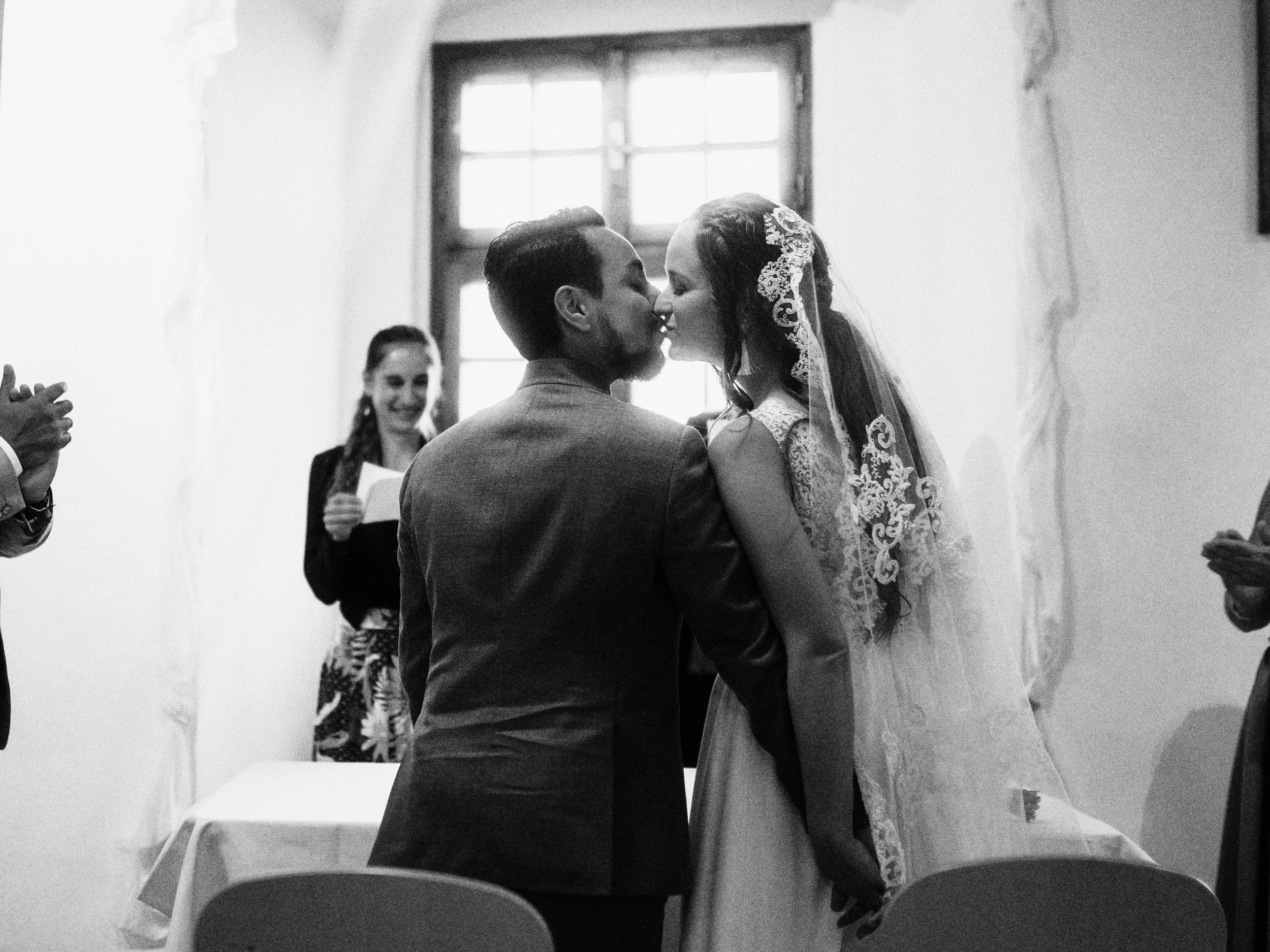 Ein Hochzeitspaar küsst sich während einer Eheschließung in einem Raum mit großem Fenster im Hintergrund, im Vordergrund und auf der Seite sind Gäste.