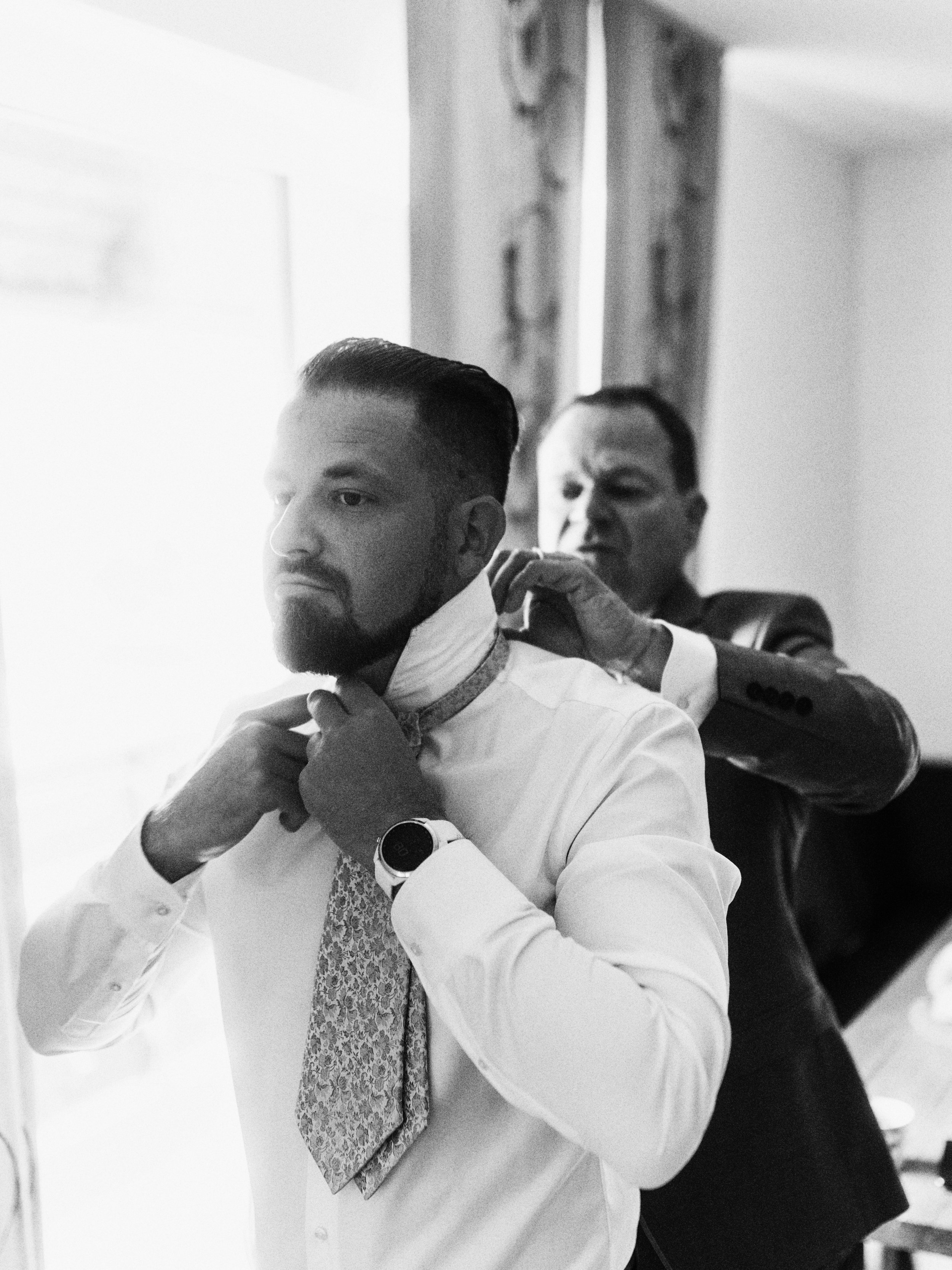 Zwei Männer, einer hilft dem anderen, seinen Hemdkragen zu richten, im Hintergrund ein Fenster und eine Wand.