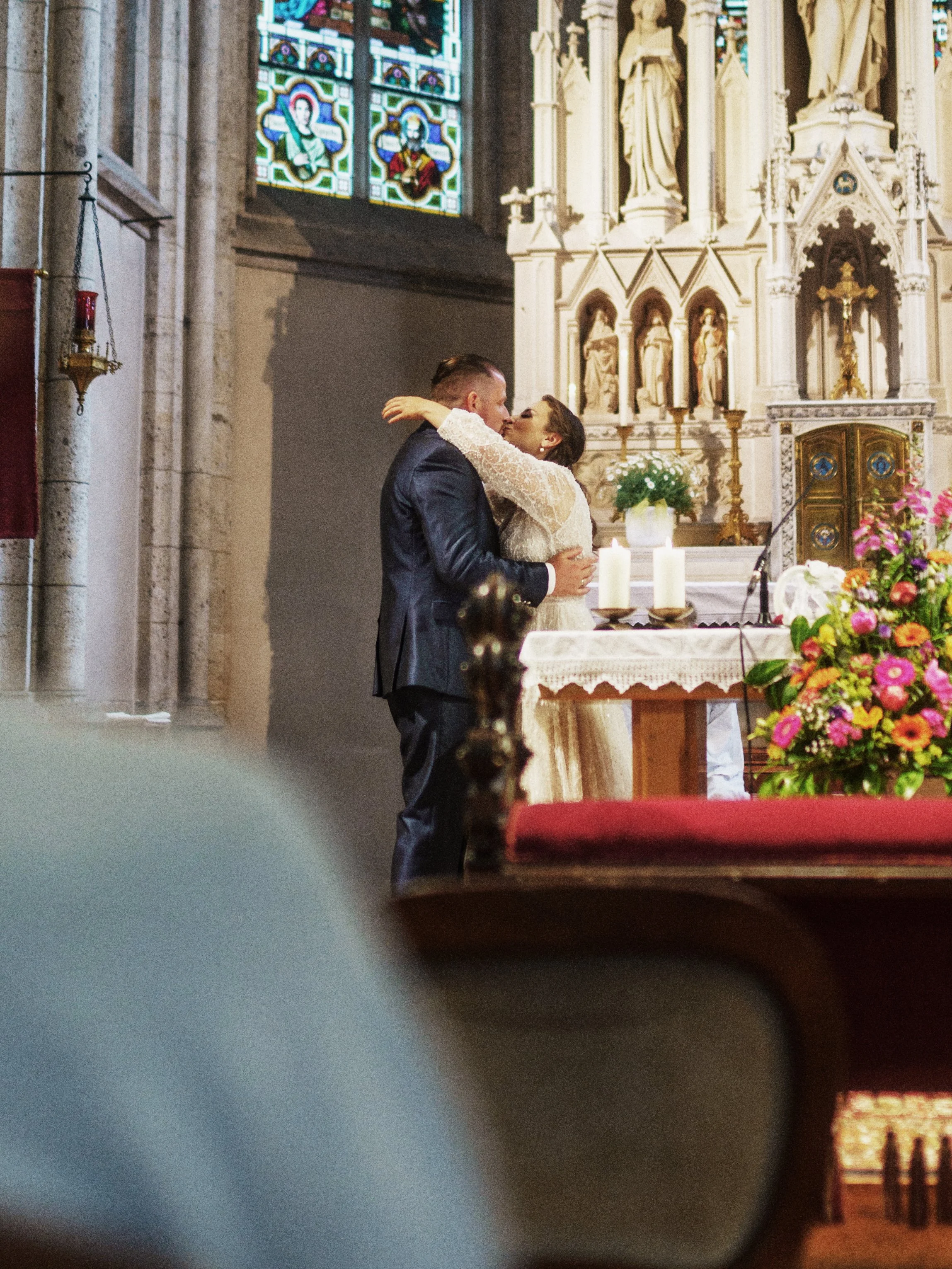 Ein Hochzeitspaar in einer Kirche, umarmend, während sie vor dem Altar stehen, mit bunten Blumen und Kerzen.