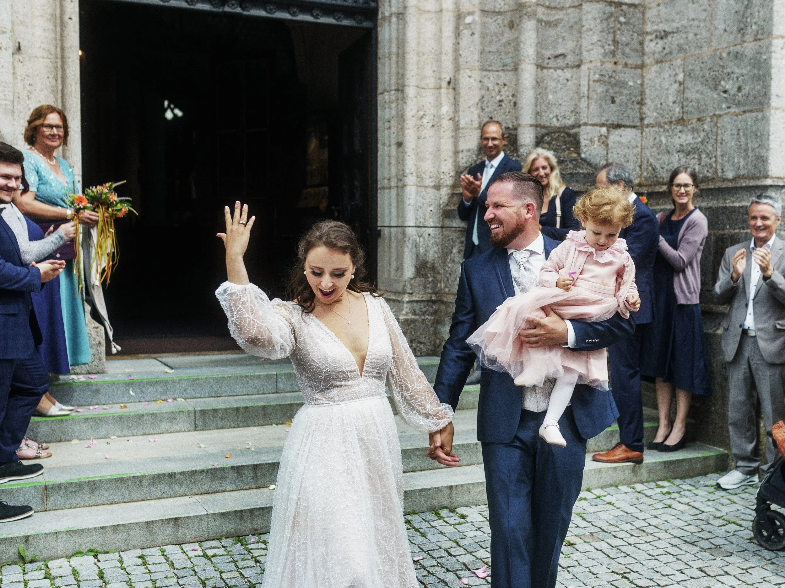 Braut und Bräutigam verlassen Kirche, umgeben von jubelnden Gästen, die applaudieren und Blumen streuen.
