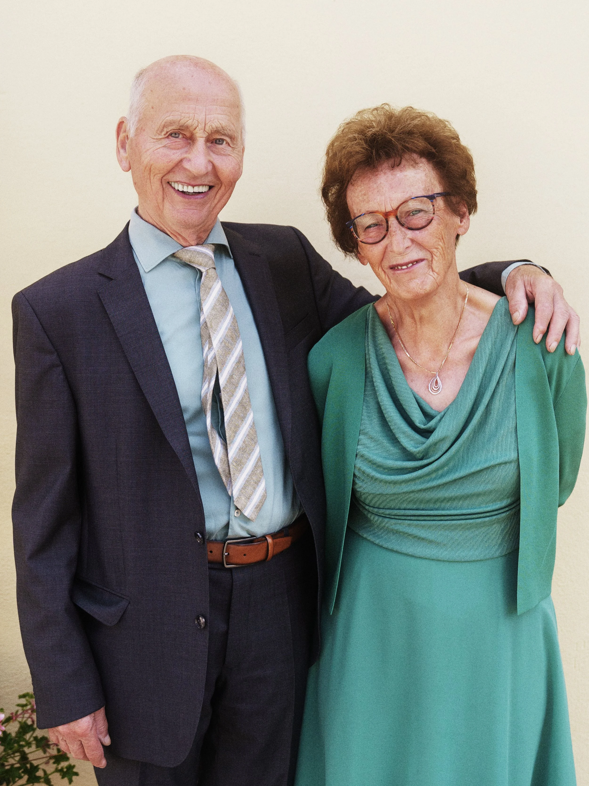 Ein älteres Paar, ein Mann in Anzug und eine Frau in einem grünen Kleid, stehen nebeneinander und lächeln, wobei der Mann einen Arm um die Schulter der Frau legt.