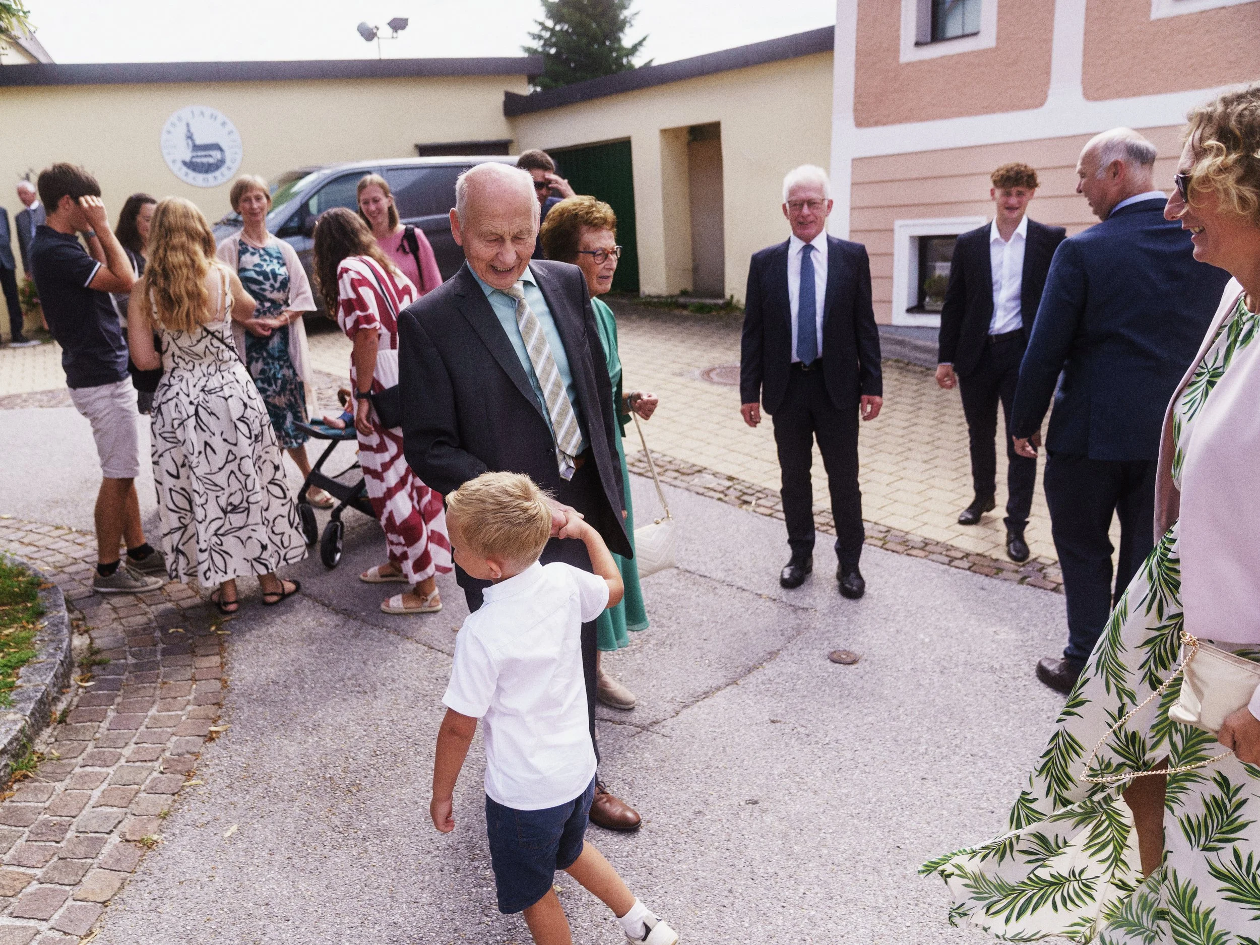 Gruppe von Menschen, überwiegend Erwachsene, die draußen stehen und sich unterhalten. Es ist eine Mischung aus Männern und Frauen, einige tragen Anzüge. Im Vordergrund ist ein älterer Mann, der lächelnd mit einem kleinen Jungen spricht.