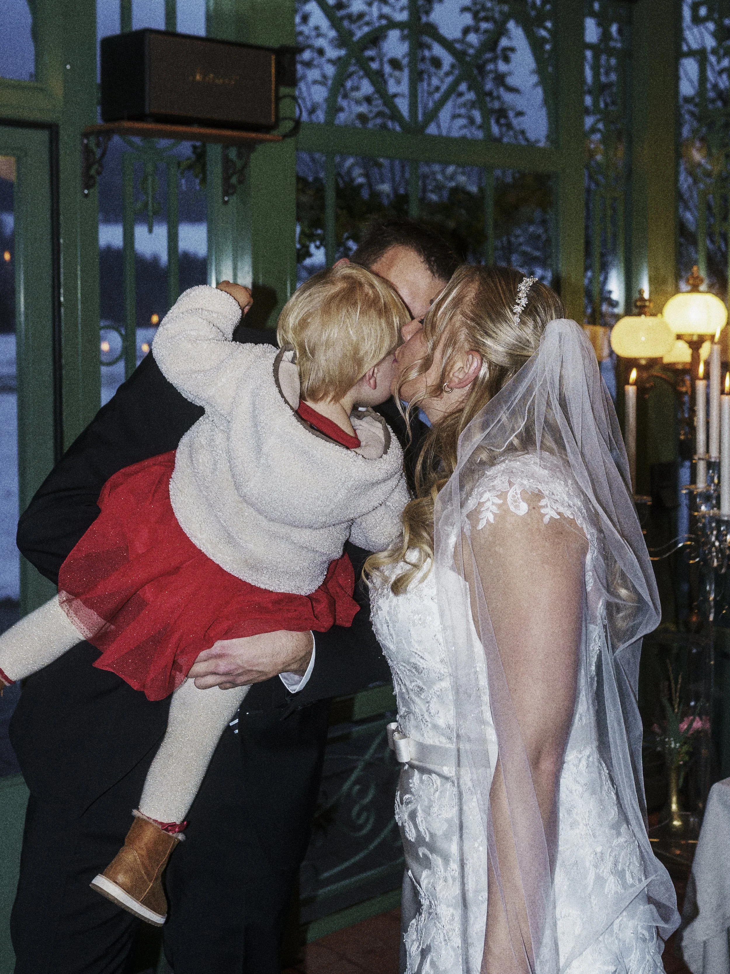 Ein Brautpaar küsst ein Kind bei einer Hochzeitsfeier in einem festlich dekorierten Raum.