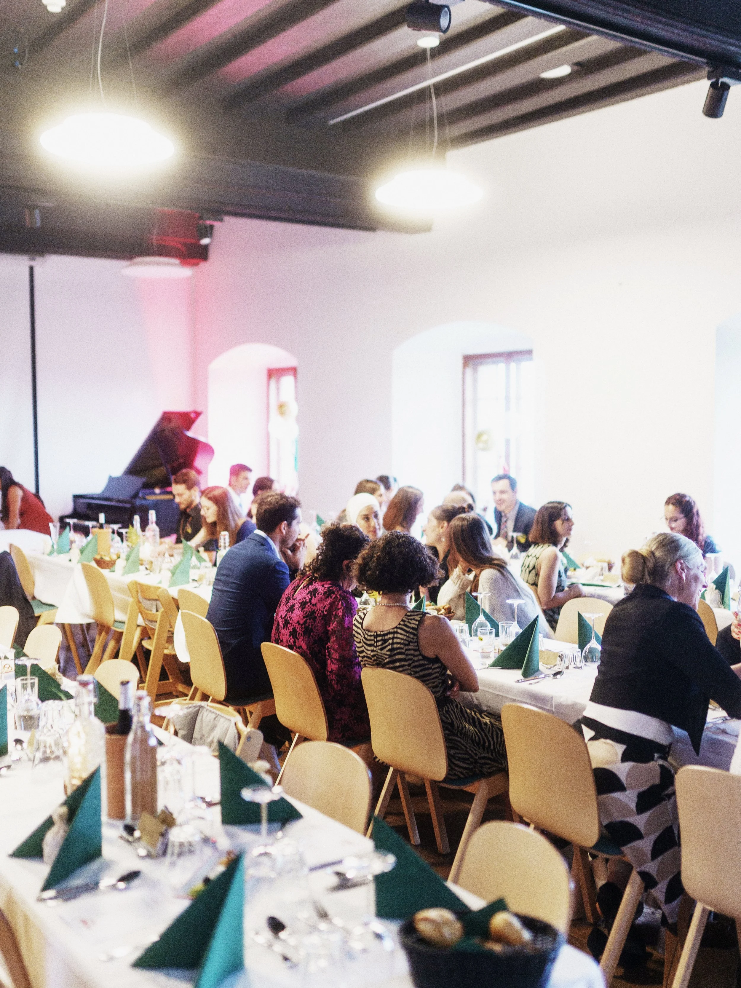 Menschen sitzen an einem festlich gedeckten langen Tisch in einem hellen Raum mit hohen Fenstern, es scheint sich um eine Feier oder einen besonderen Anlass zu handeln.
