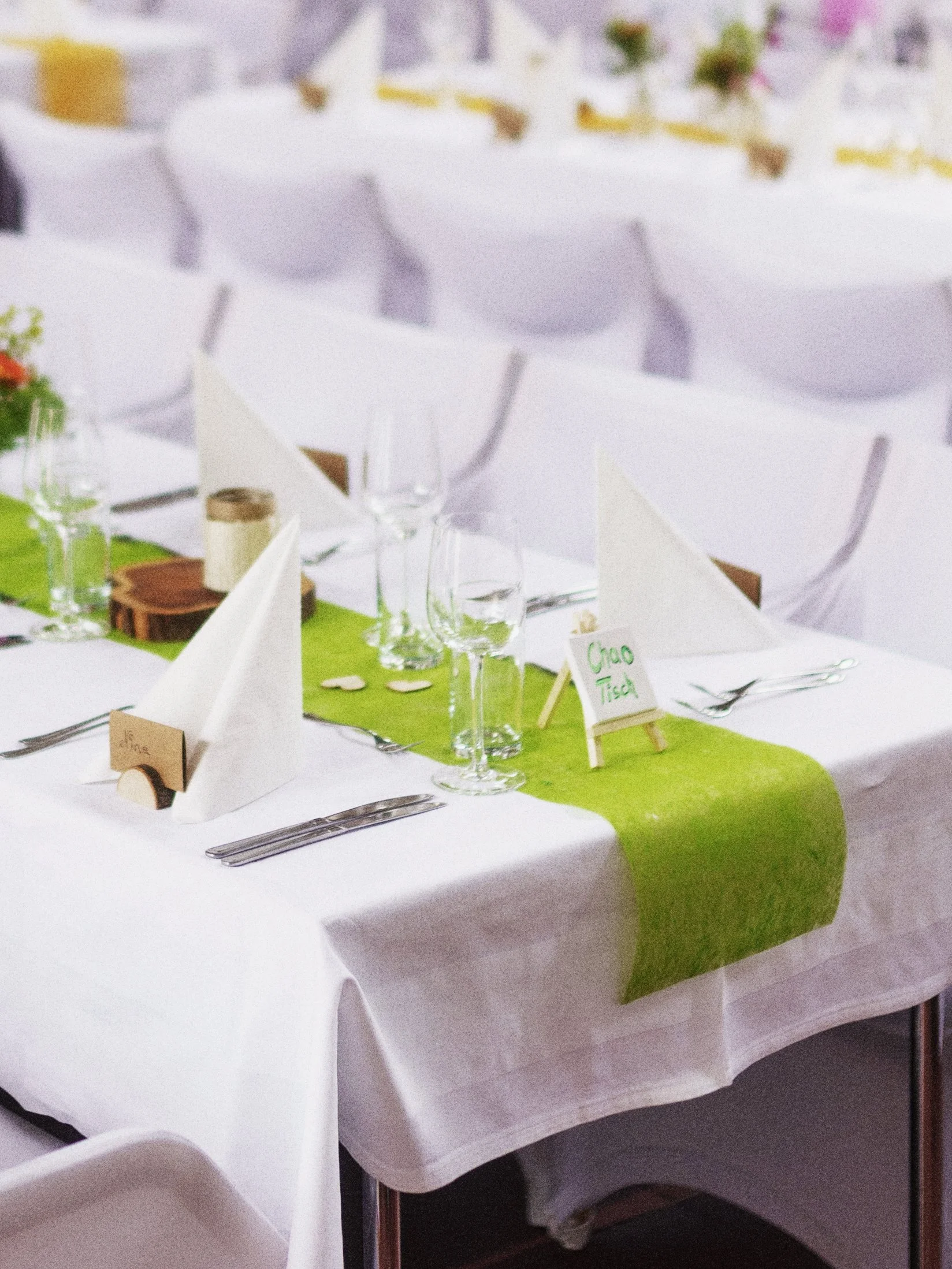 Festlich gedeckter Tisch mit weißen Tischtüchern, einer grünen Tischdecke, Gläsern, Besteck, gefalteten weißen Servietten, kleinen Platzkarten und Dekorationen für eine Feier.