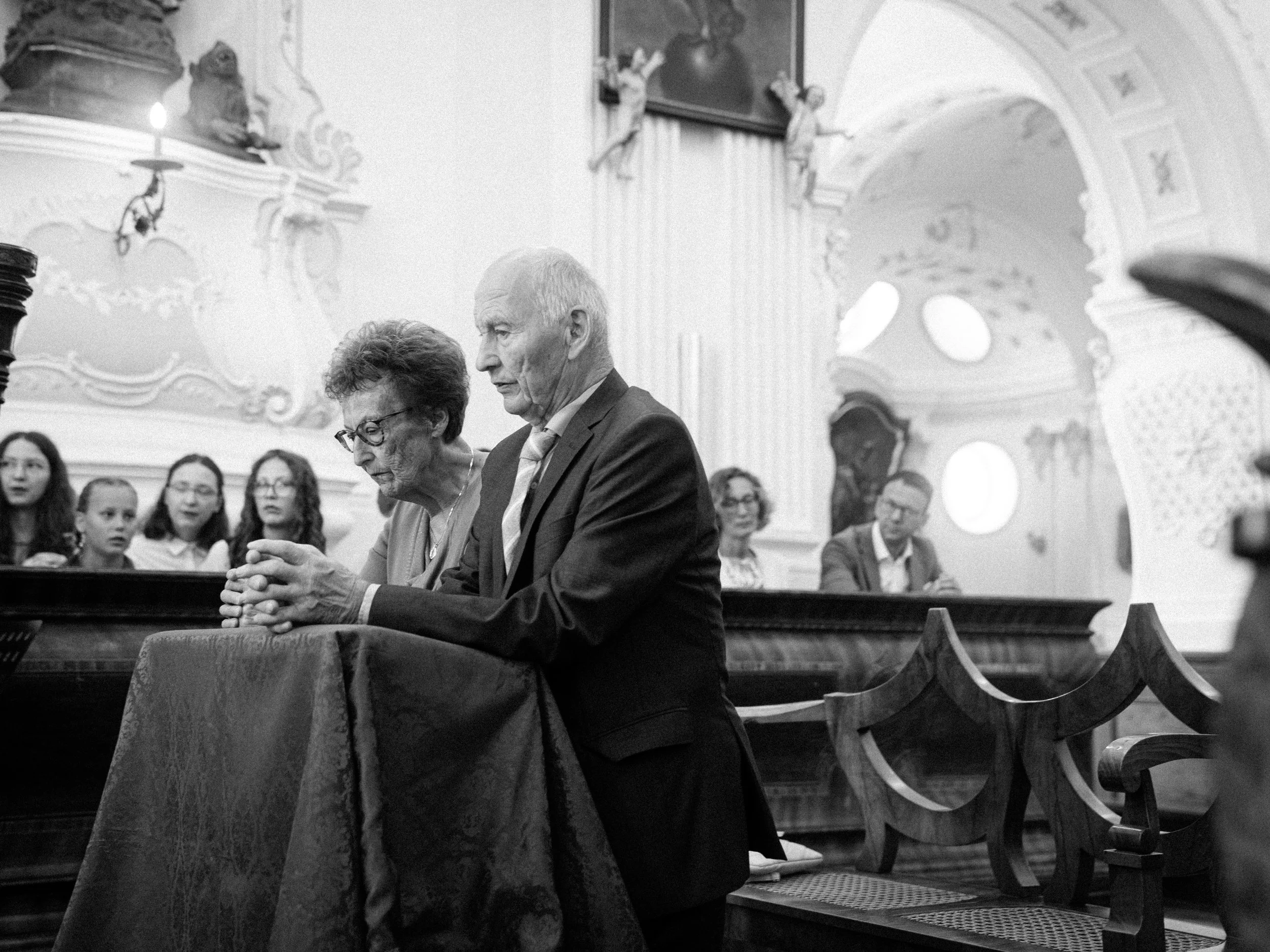 Älteres Paar betet in einer Kirche während eines Gottesdienstes, umgeben von Gläubigen, mit kunstvoller Barockarchitektur im Hintergrund.