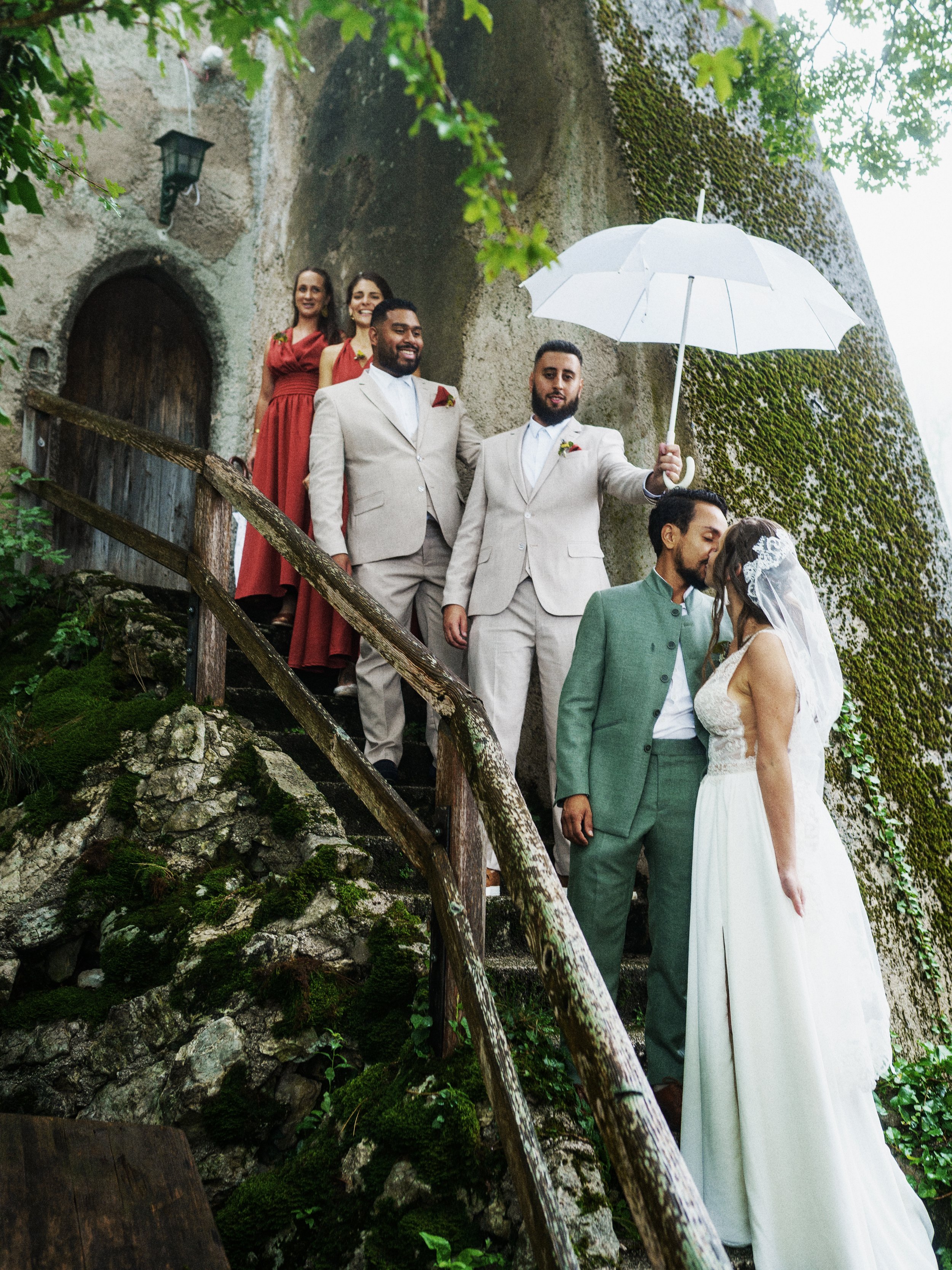 Ein Brautpaar küsst sich, während der Bräutigam einen weißen Regenschirm über die Braut hält. Dahinter stehen Trauzeugen und Brautjungfern, auf einer Stein- und Holztreppe vor einem alten, mit Moos bewachsenen Steinbau.
