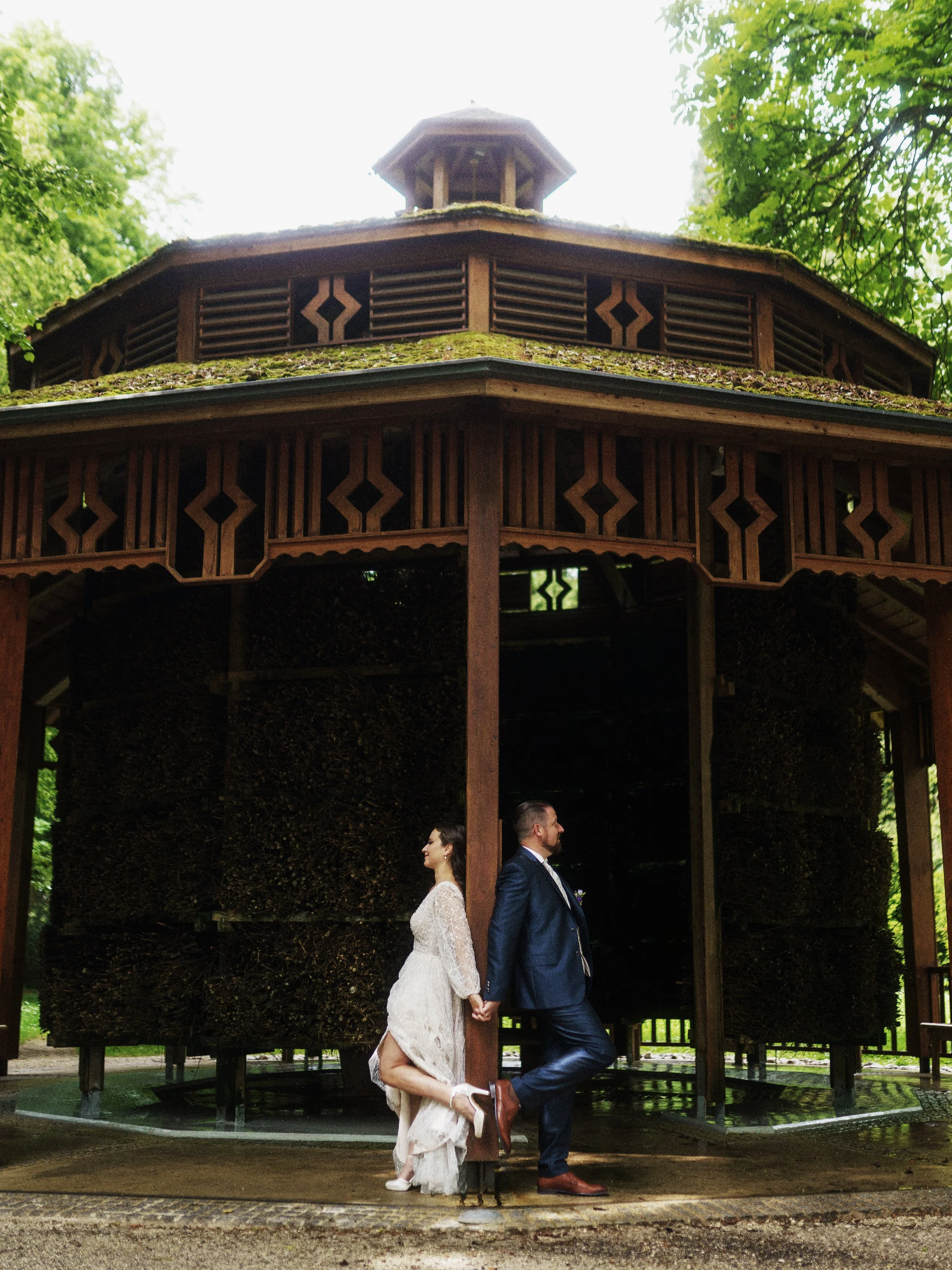 Ein Brautpaar steht in einem Holz-Pavillon, der von außen sichtbar ist und trägt formelle Kleidung. Die Frau trägt ein weißes Brautkleid, der Mann einen dunklen Anzug.