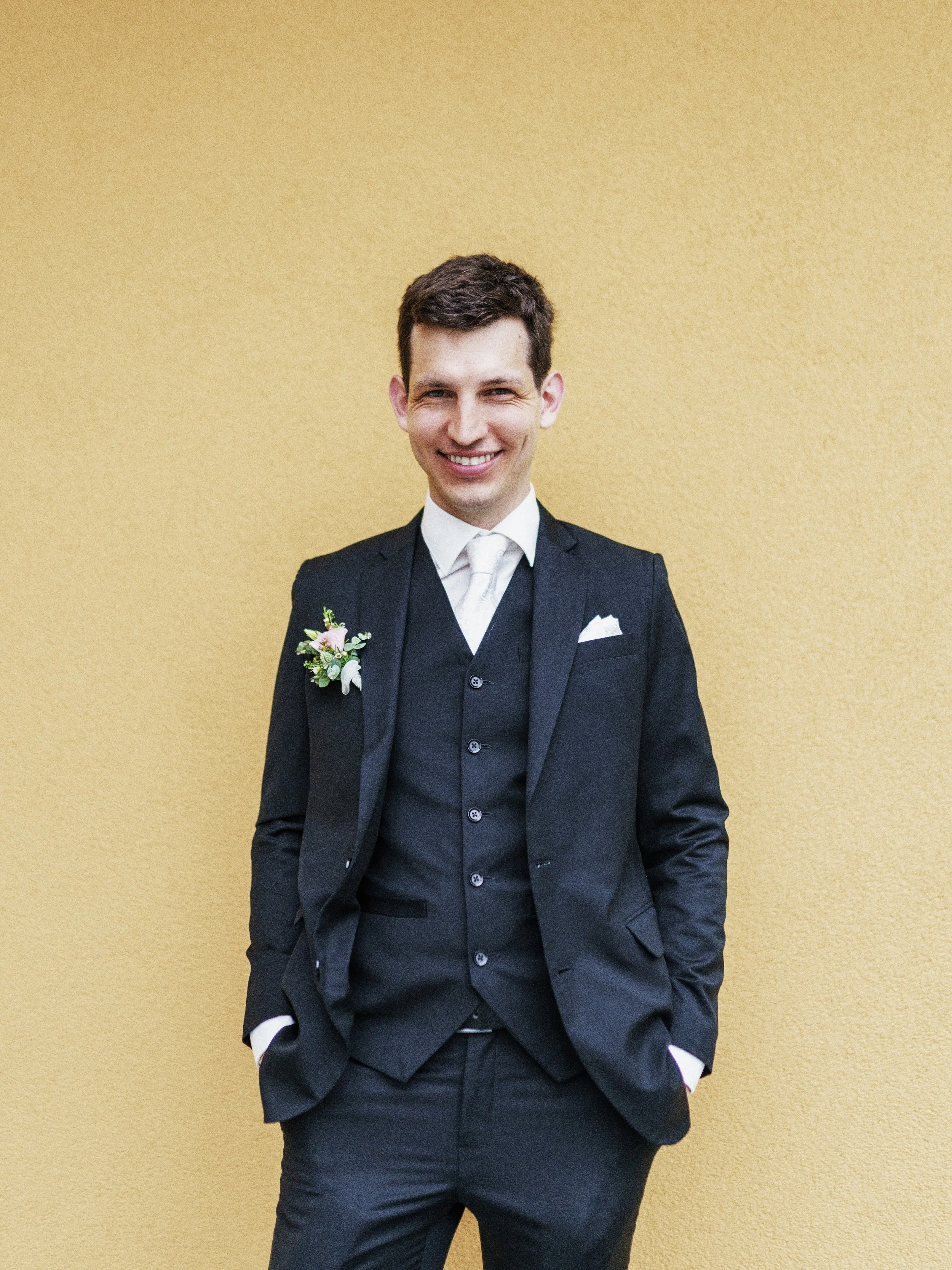 Ein junger Mann in formeller Kleidung, mit Anzug und Krawatte, steht vor einer gelben Wand und lächelt in die Kamera.