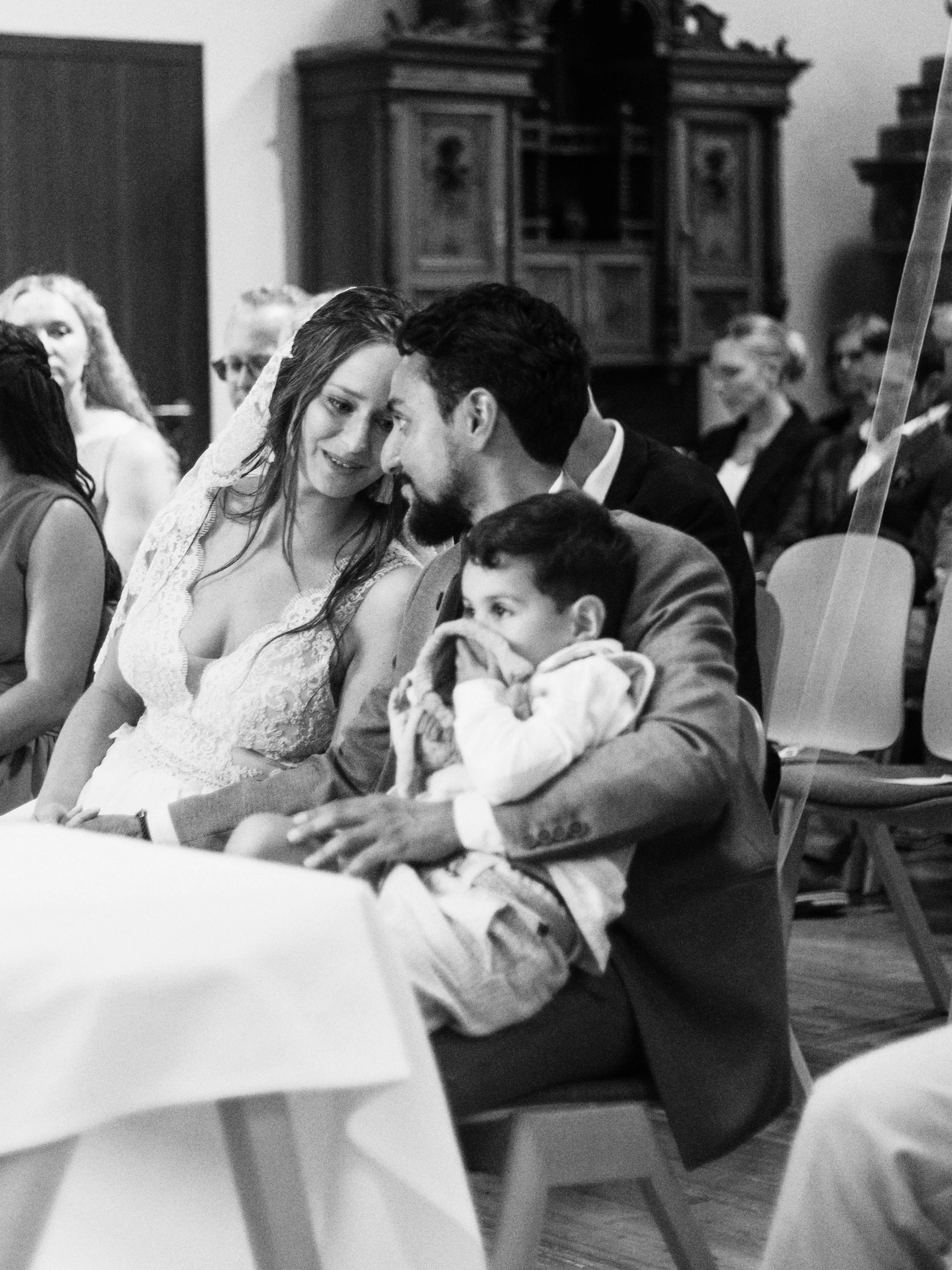 Eine schwarze und weiße Fotografie eines Hochzeitsfeier mit einer Braut, einem Bräutigam, einem kleinen Kind und weiteren Gästen im Hintergrund.