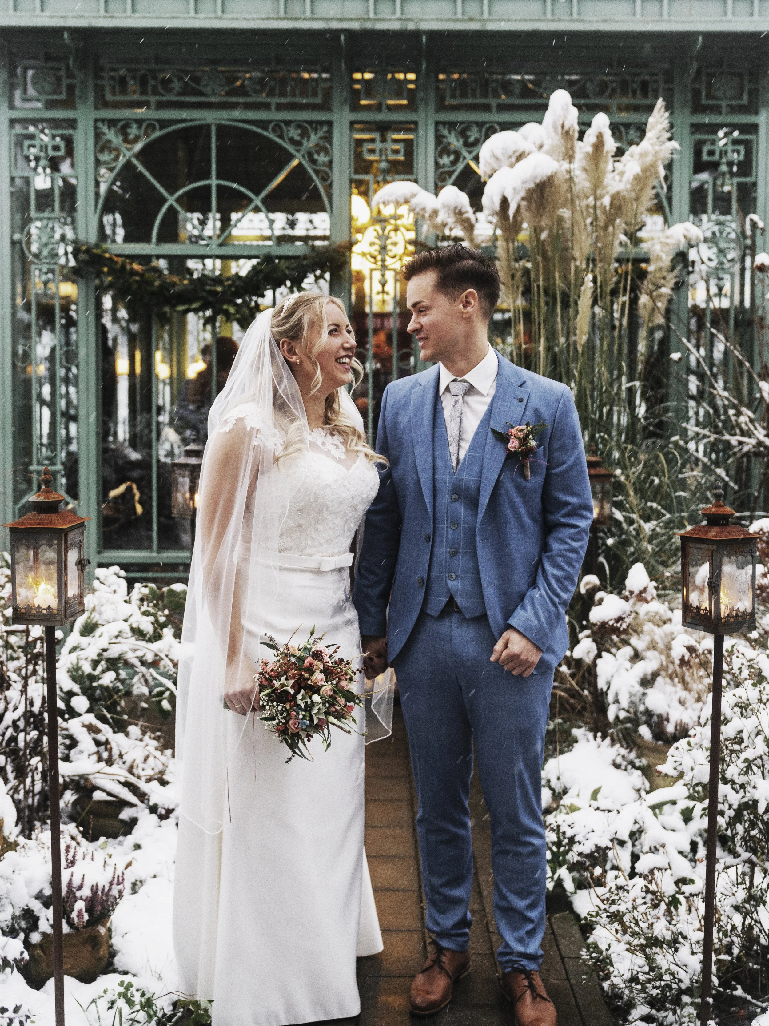 Verheiratetes Paar steht im Schnee vor einem gläsernen, grünen Garten im Winter, hält Händchen, lächelt und schaut sich liebevoll an, die Braut trägt ein weißes Brautkleid und Schleier, die Tasche mit Blumen, das Paar ist umgeben von Schneebedeckten Pflanzen und Laternen.