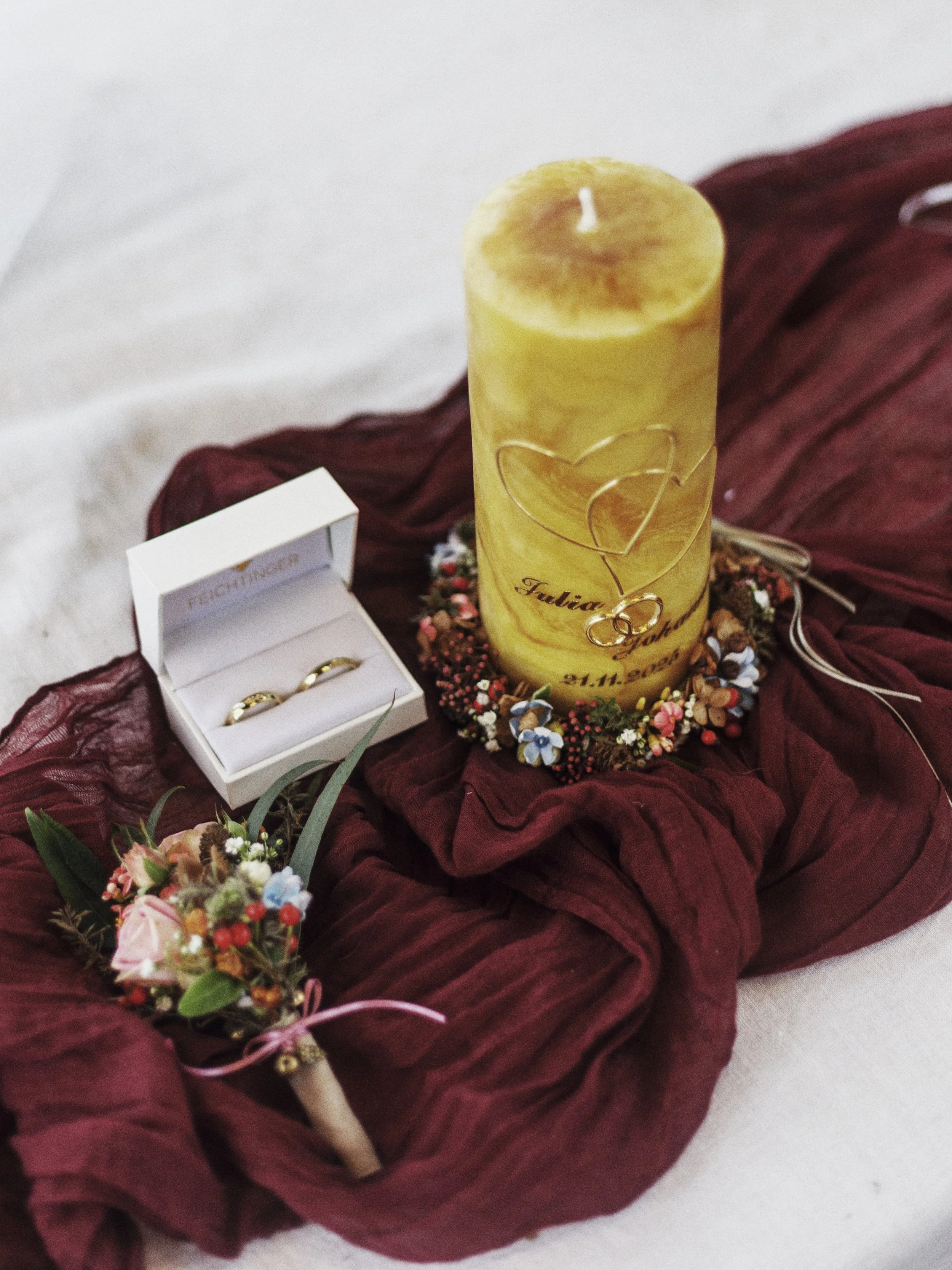 Ein gelber Kerze mit Herzanhänger, eine kleine Schachtel mit Eheringen, und Blumenarrangements auf roten Stoff, vermutlich für eine Hochzeit.