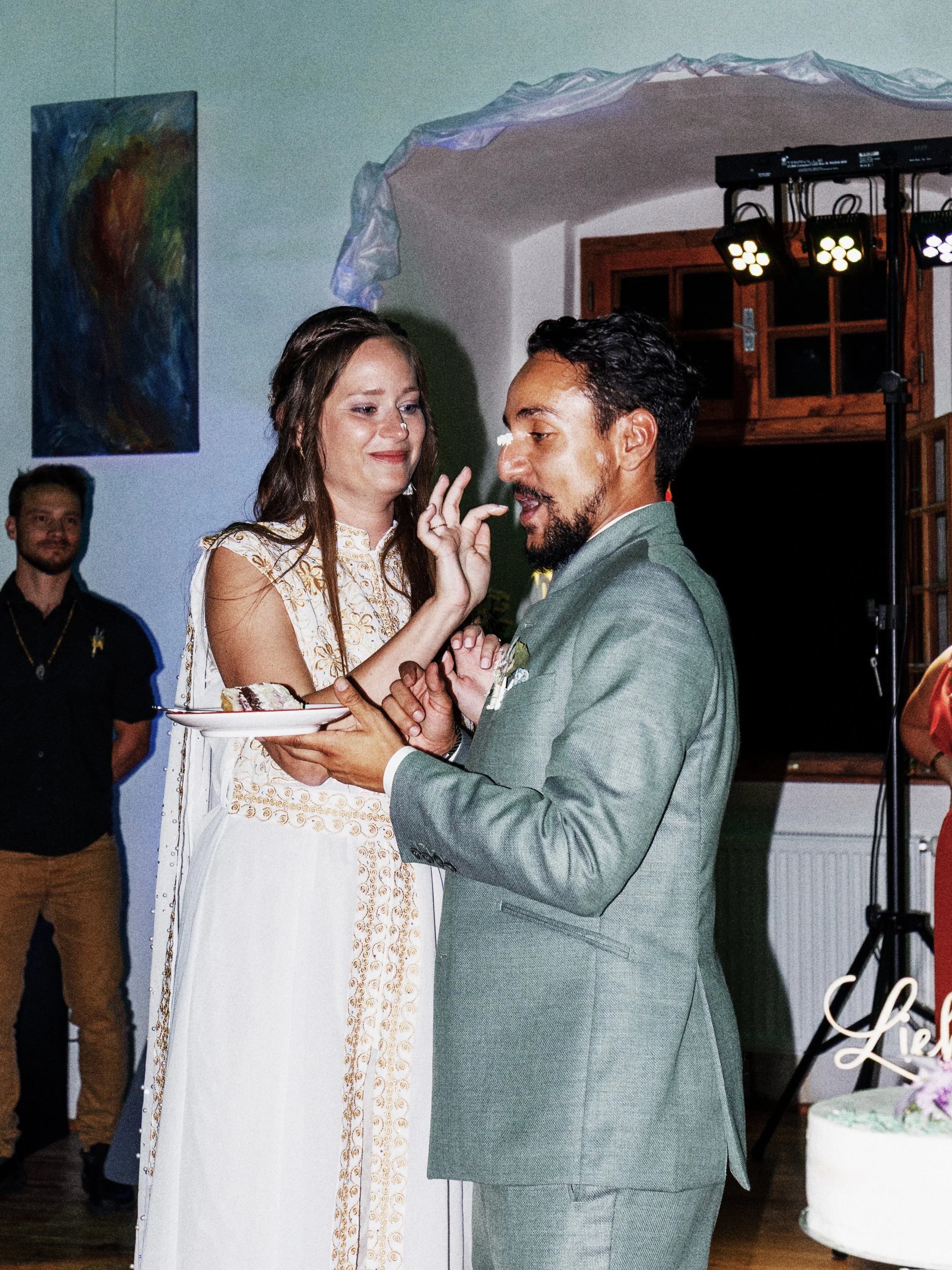 Ein Brautpaar bei einer Hochzeit, die Braut in einem weißen Kleid mit goldenen Details, der Bräutigam in einem grauen Anzug. Sie tauschen ein Stück Hochzeitstorte aus, im Hintergrund sind weitere Gäste zu sehen, und es gibt bunte Beleuchtung.