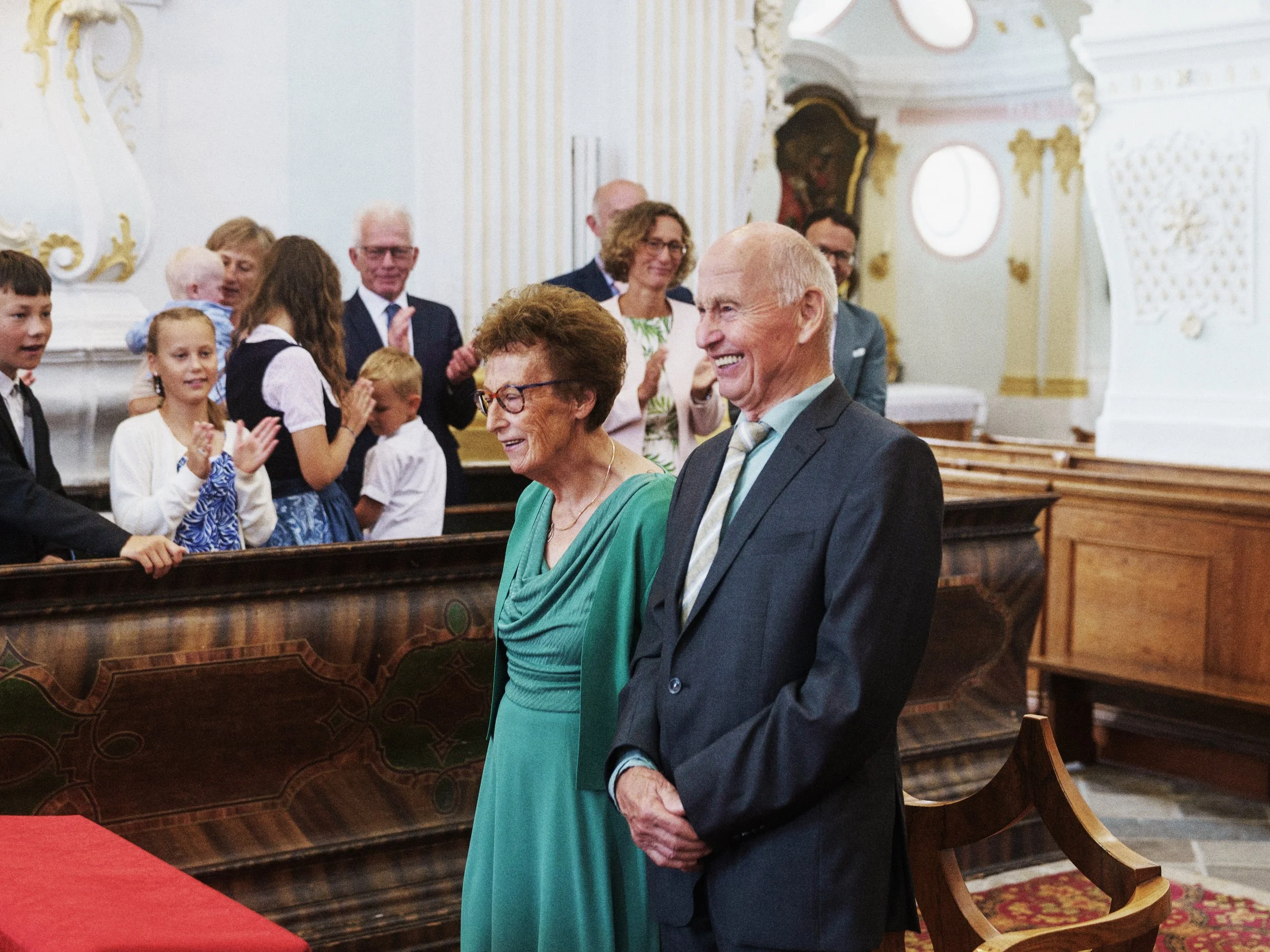 Ein älteres Paar, ein Mann und eine Frau, stehen in einer Kirche, lächeln und sind bei einer Zeremonie oder Feier aufgeregt. Im Hintergrund sind weitere Menschen, Kinder und Erwachsene, die applaudieren und sich freuen.