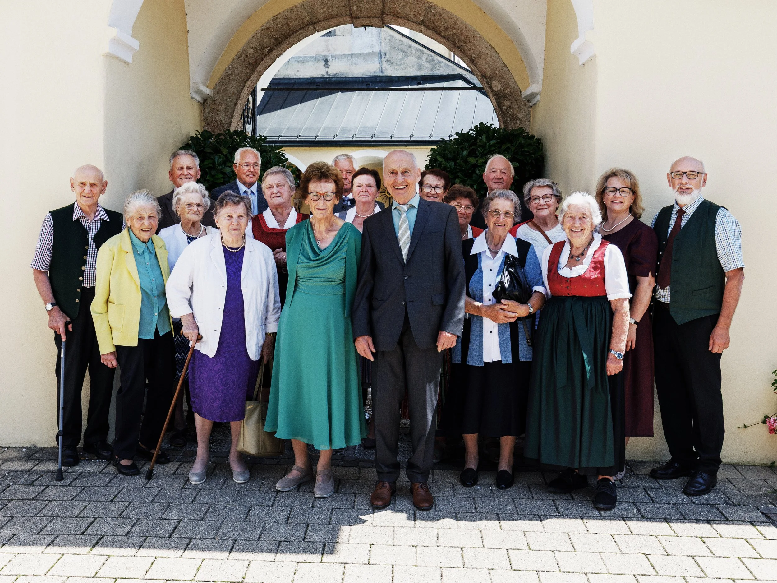 Gruppe älterer Menschen, hauptsächlich Frauen, pose in traditioneller Tracht und formeller Kleidung vor einem historischen Gebäude mit Bogen im Hintergrund.