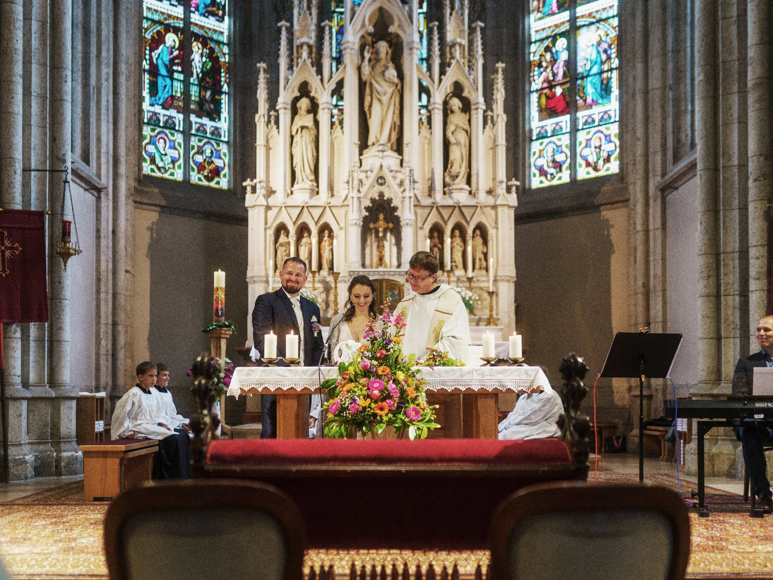 Ein Hochzeitspaar steht zusammen mit einem Priester an einem Altar in einer gotischen Kirche. Es gibt bunte Blumen, Kerzen und Bänke, auf denen zwei Messdiener sitzen. Bunte Kirchenfenster sind im Hintergrund sichtbar.