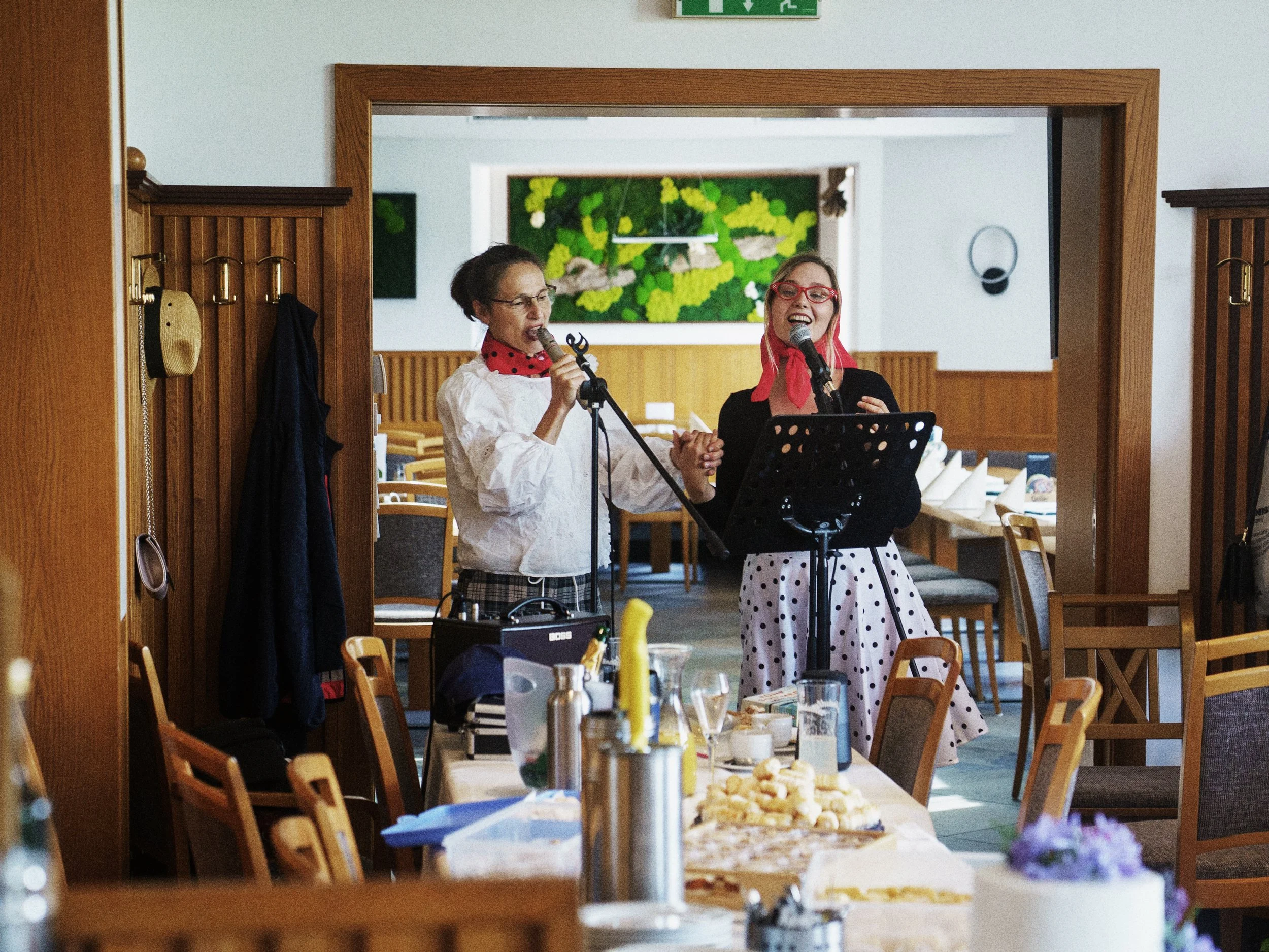 Zwei Frauen singen in einem Restaurant, mit Mikrofonen, Tischen mit Essen und Getränken im Vordergrund.