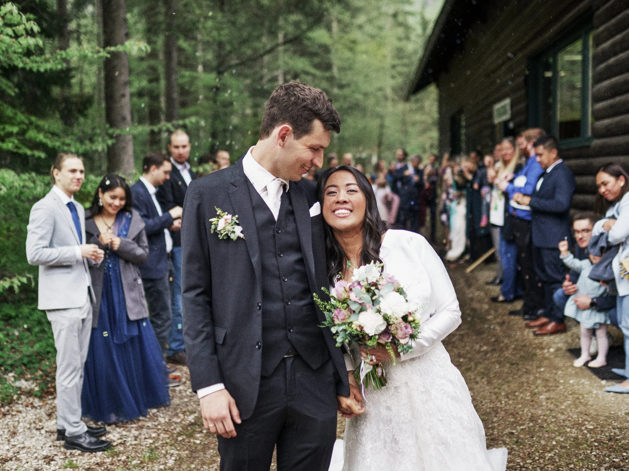 Ein heiratswilliges Paar, ein Mann in einem dunklen Anzug und eine Frau in einem weißen Brautkleid, steht Hand in Hand im Freien, umgeben von Freunden und Familie bei einer Hochzeit im Wald.