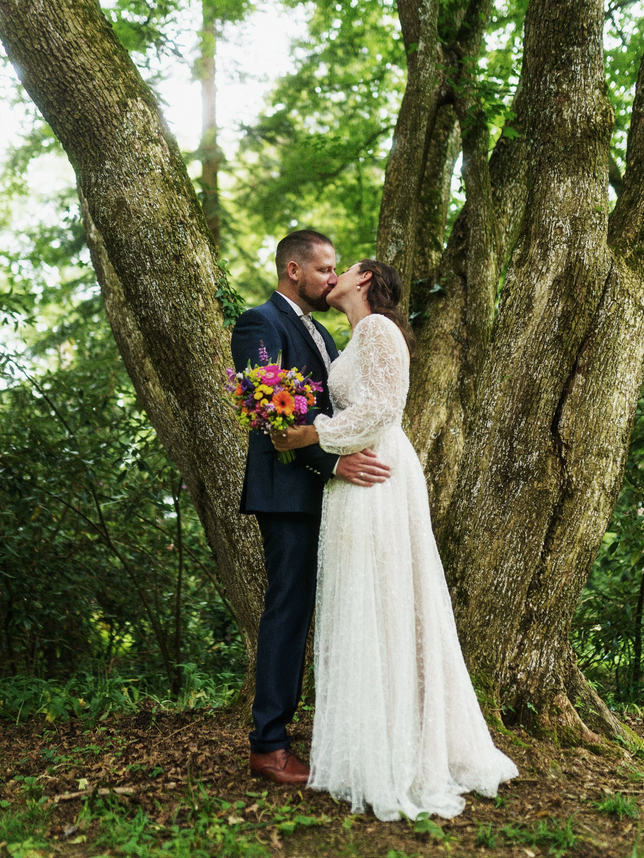 Ein Brautpaar umarmt sich und küsst sich unter einem Baum in einem grünen Wald, die Braut hält einen bunten Blumenstrauß.