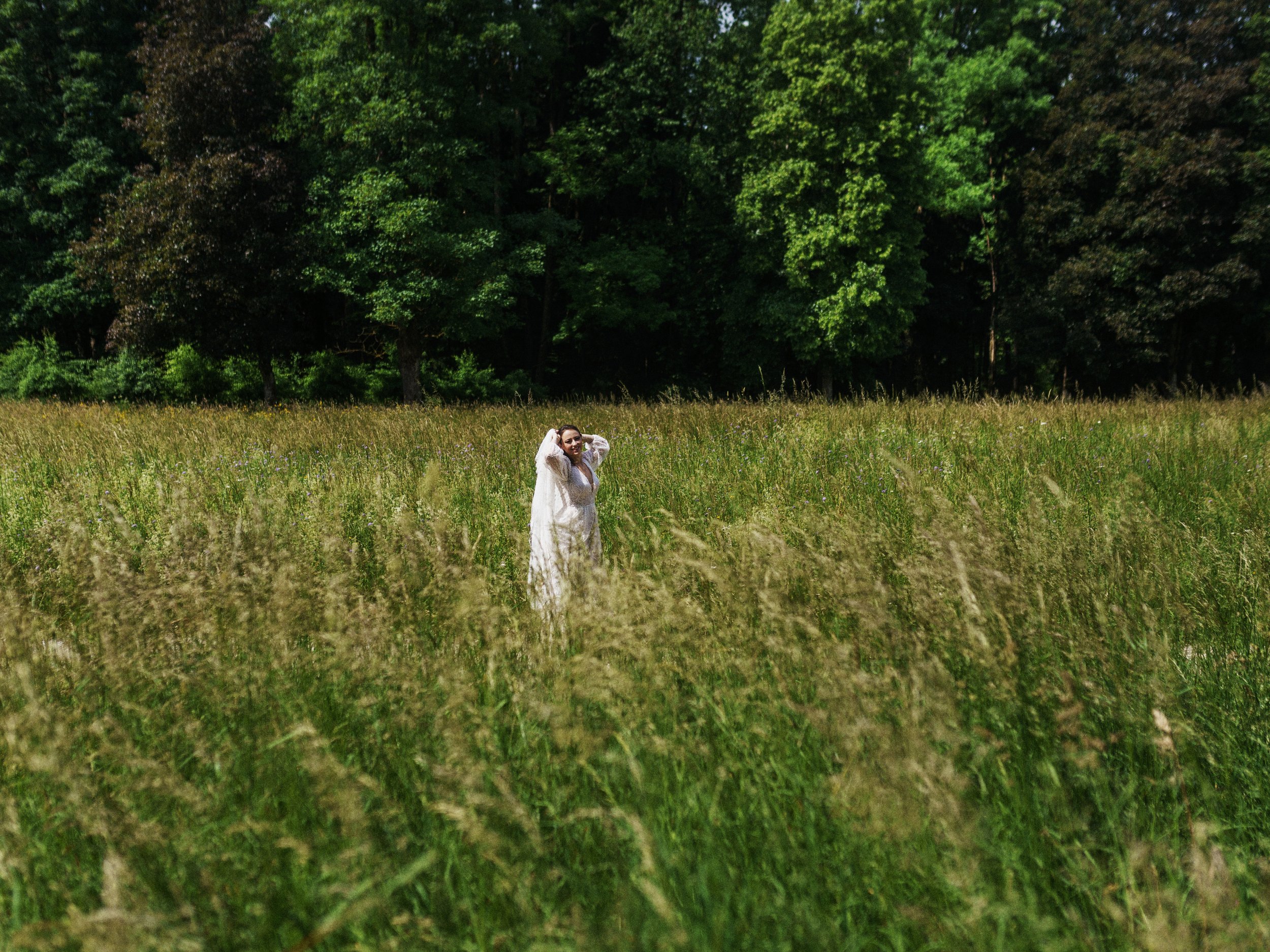 Frau in weißem Kleid steht in einem grünen Feld mit Bäumen im Hintergrund.